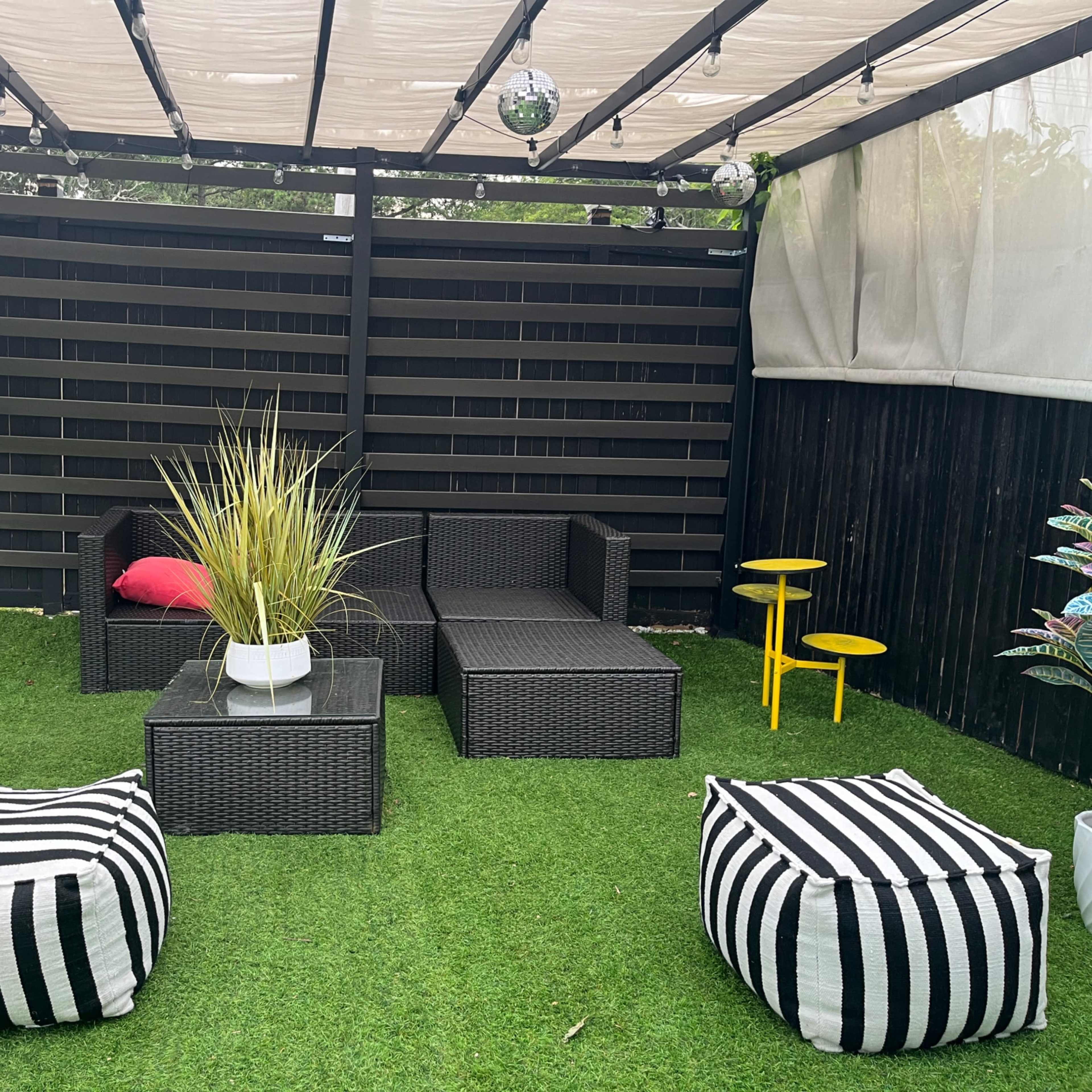 A shaded outdoor seating area featuring wicker furniture, a decorative plant, and striped poufs on artificial grass.