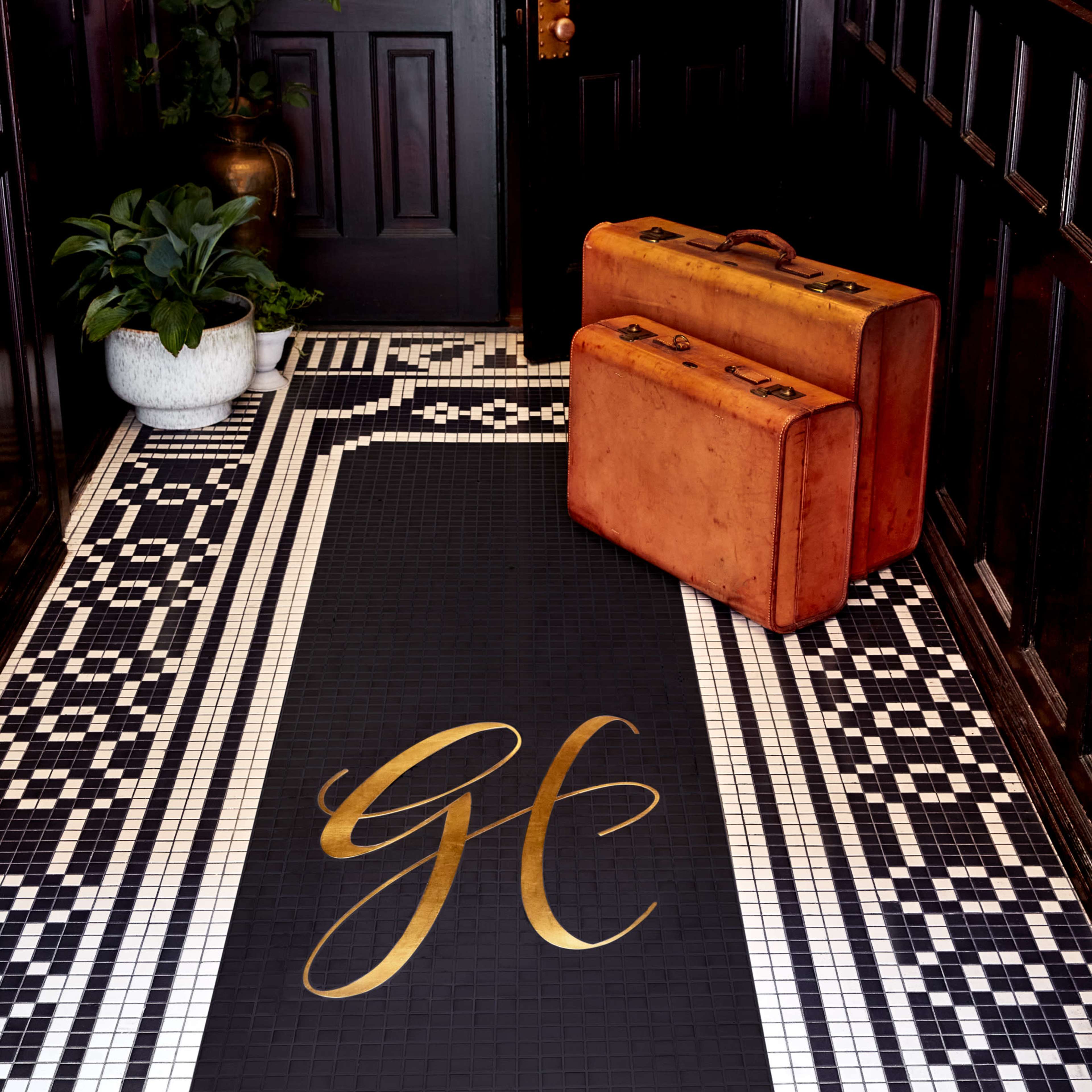 An entryway features a black mat with gold lettering prominently displaying "GE," alongside two vintage orange suitcases and a potted plant.