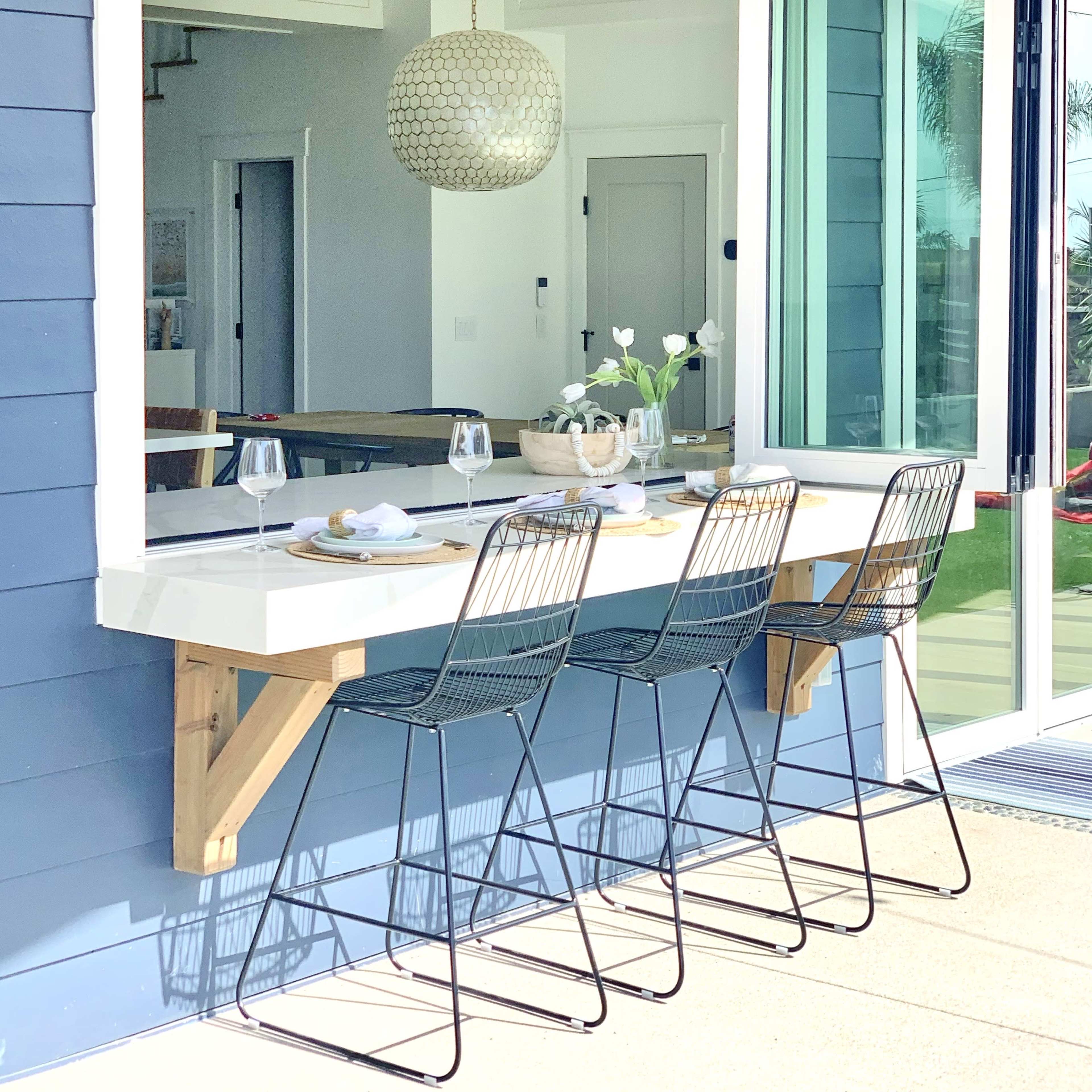 A outdoor countertop with three black metal stools in front of a large window, featuring a pendant light and a vase of flowers on the countertop.