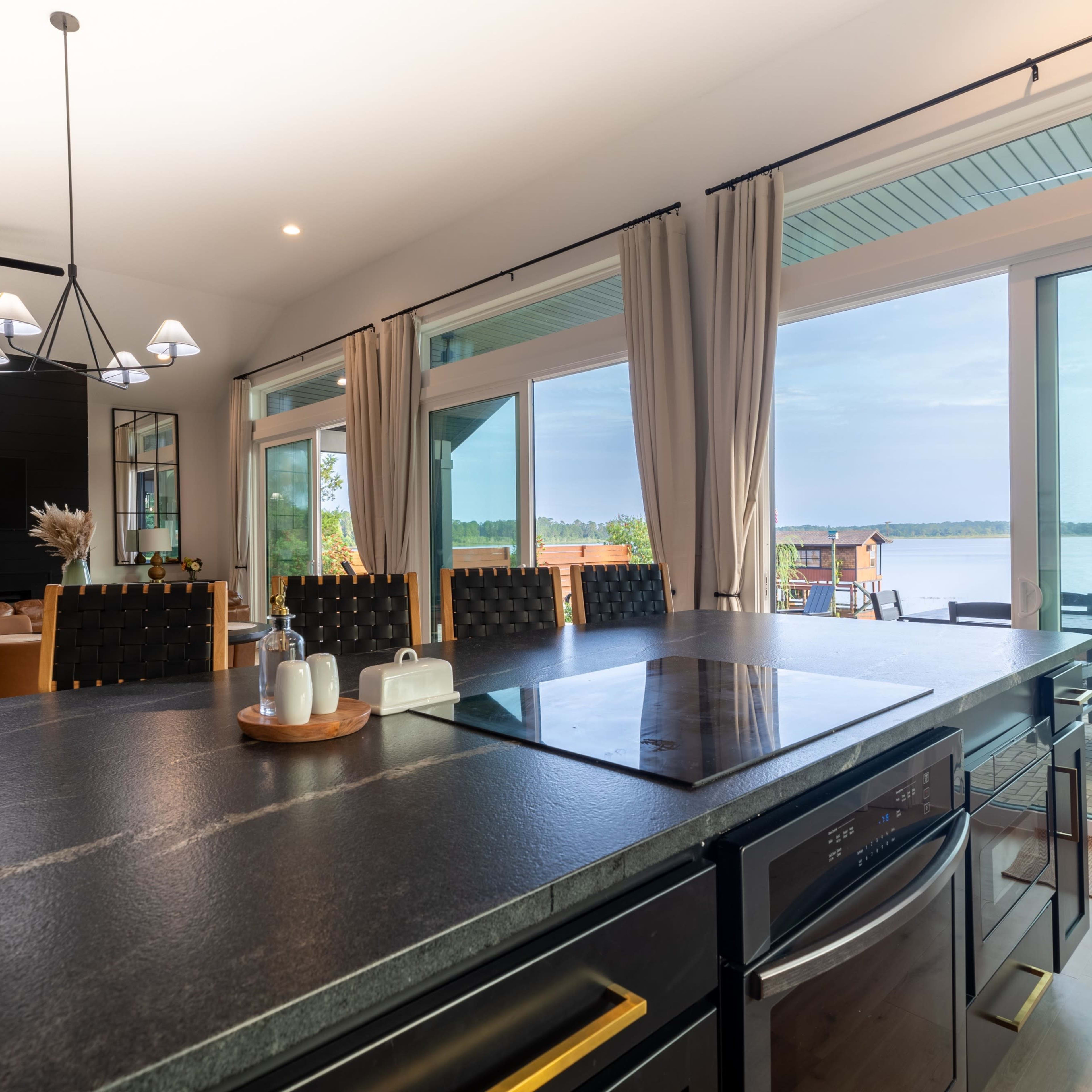 The kitchen features a dark countertop with a cooking surface, overlooking large glass doors that lead to a deck by the water.