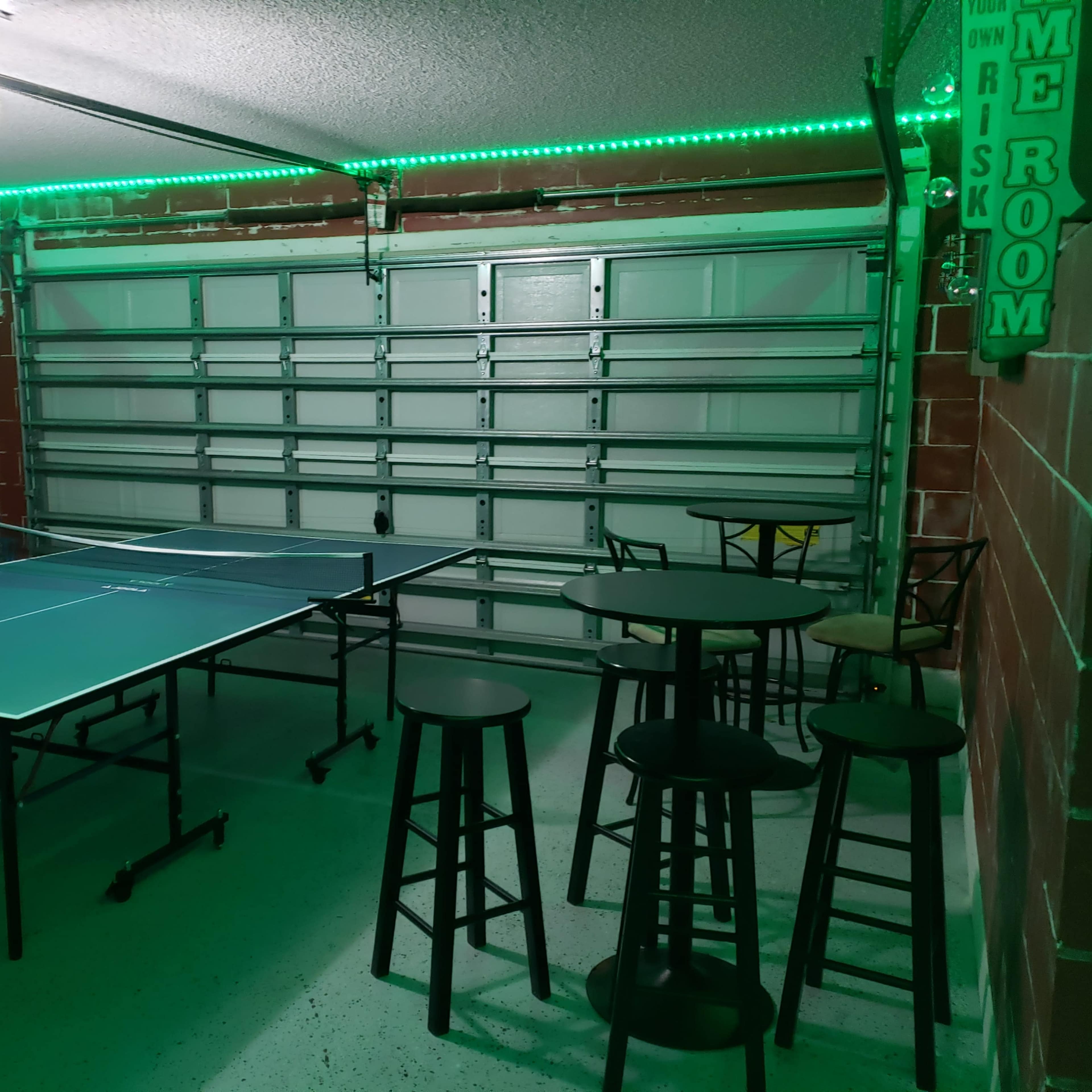 A ping pong table is set up in a garage adorned with green LED lighting and accompanied by several high stools and tables.