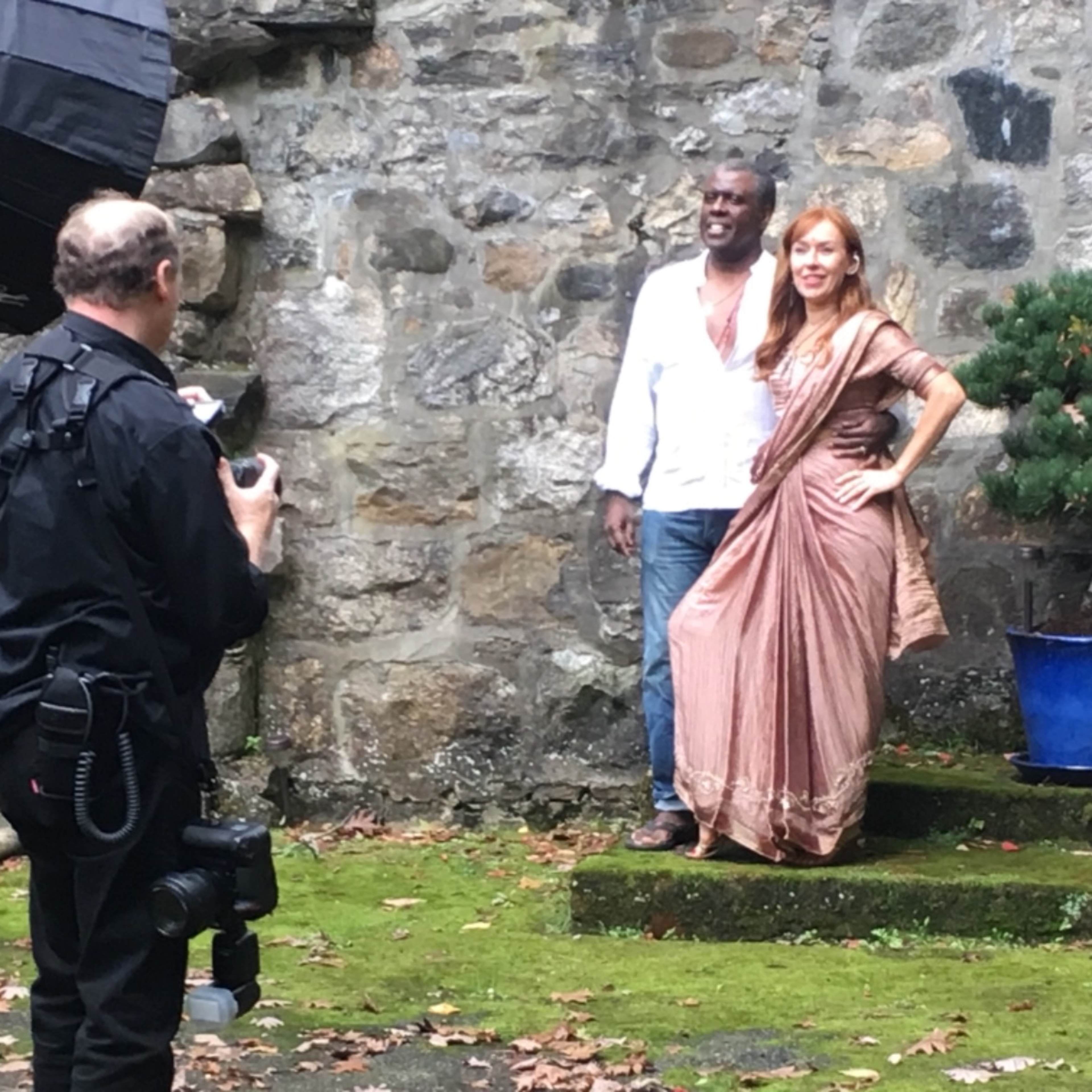 A photographer captures a couple posing in traditional attire in front of a stone wall and greenery.