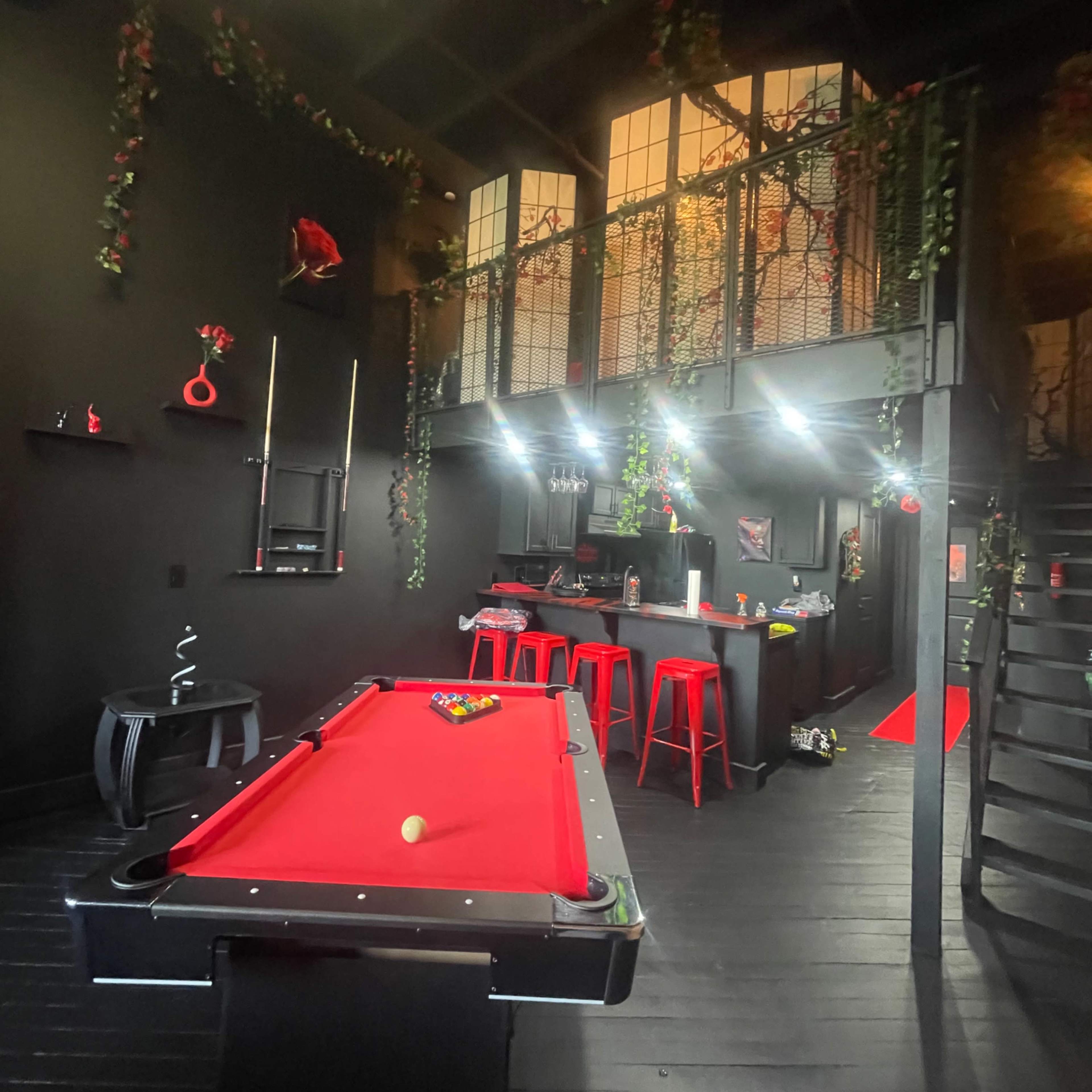 A modern game room featuring a red pool table, black walls, and a staircase leading to an upper level adorned with decorative greenery.