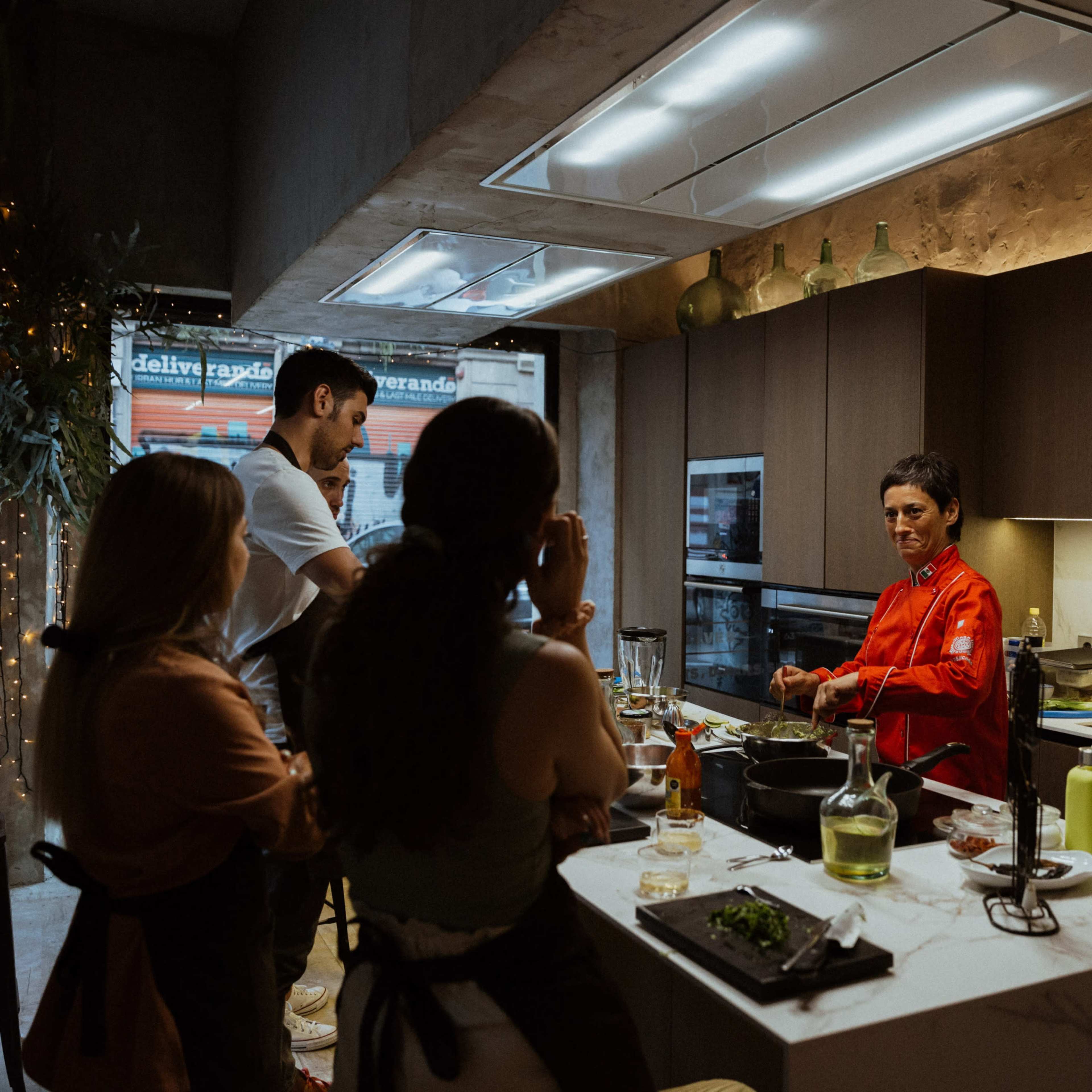 A group of four people watches as a chef in a red coat prepares food in a modern kitchen.