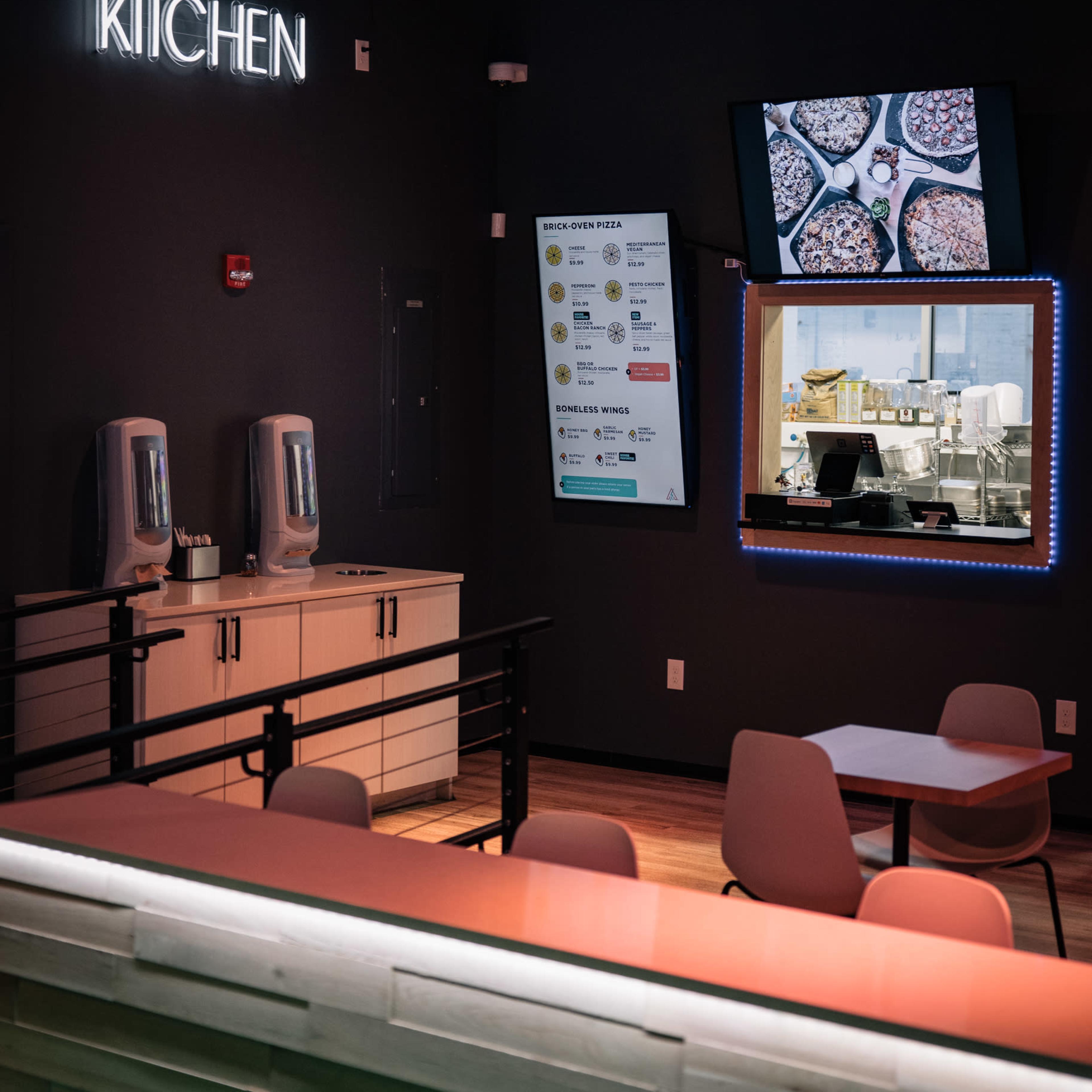 A contemporary kitchen area featuring a neon sign labeled "KITCHEN," a digital menu display, and a mirror reflecting another workspace.