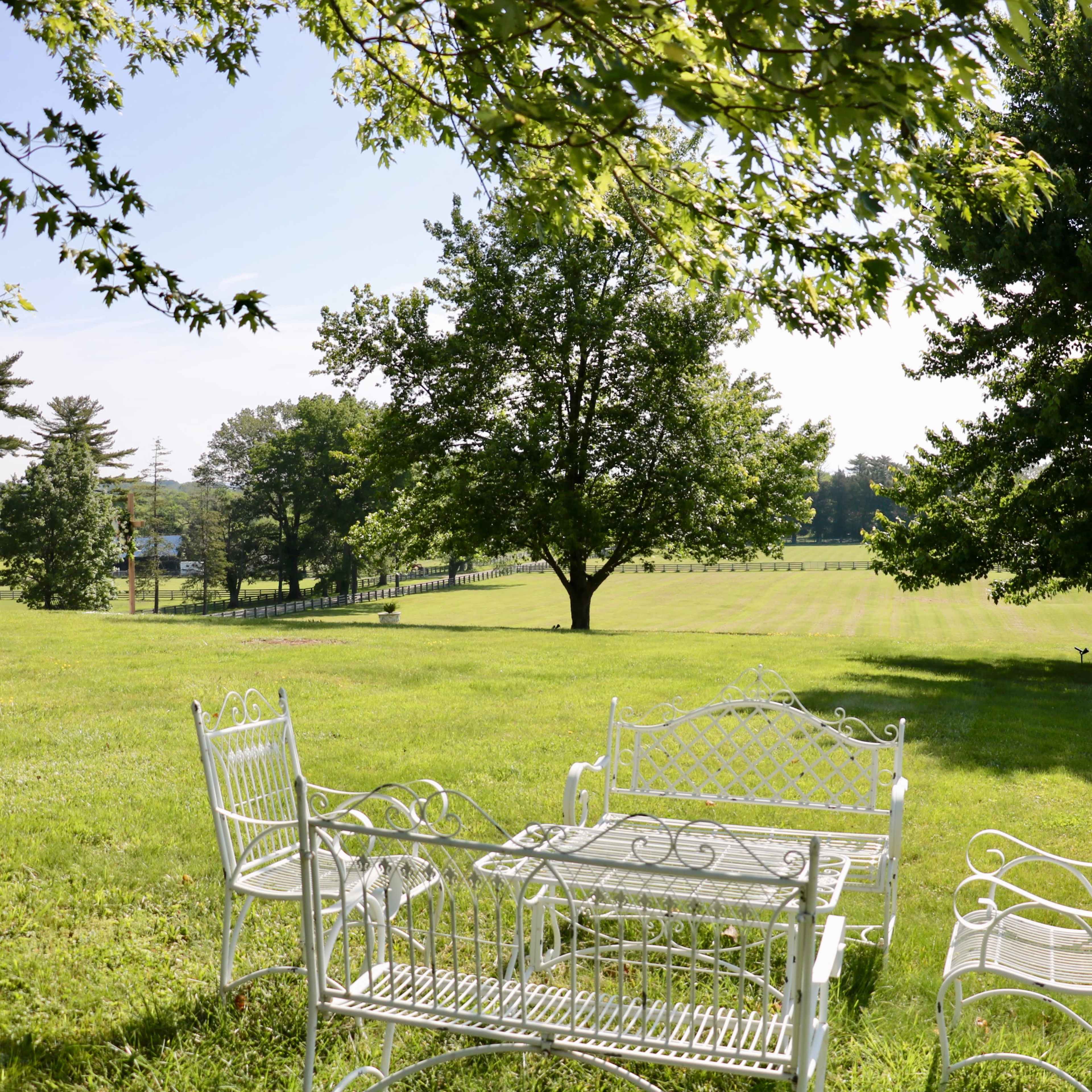 A white metal table and chairs are situated on a grassy lawn beneath two trees, with a view of rolling hills in the background.