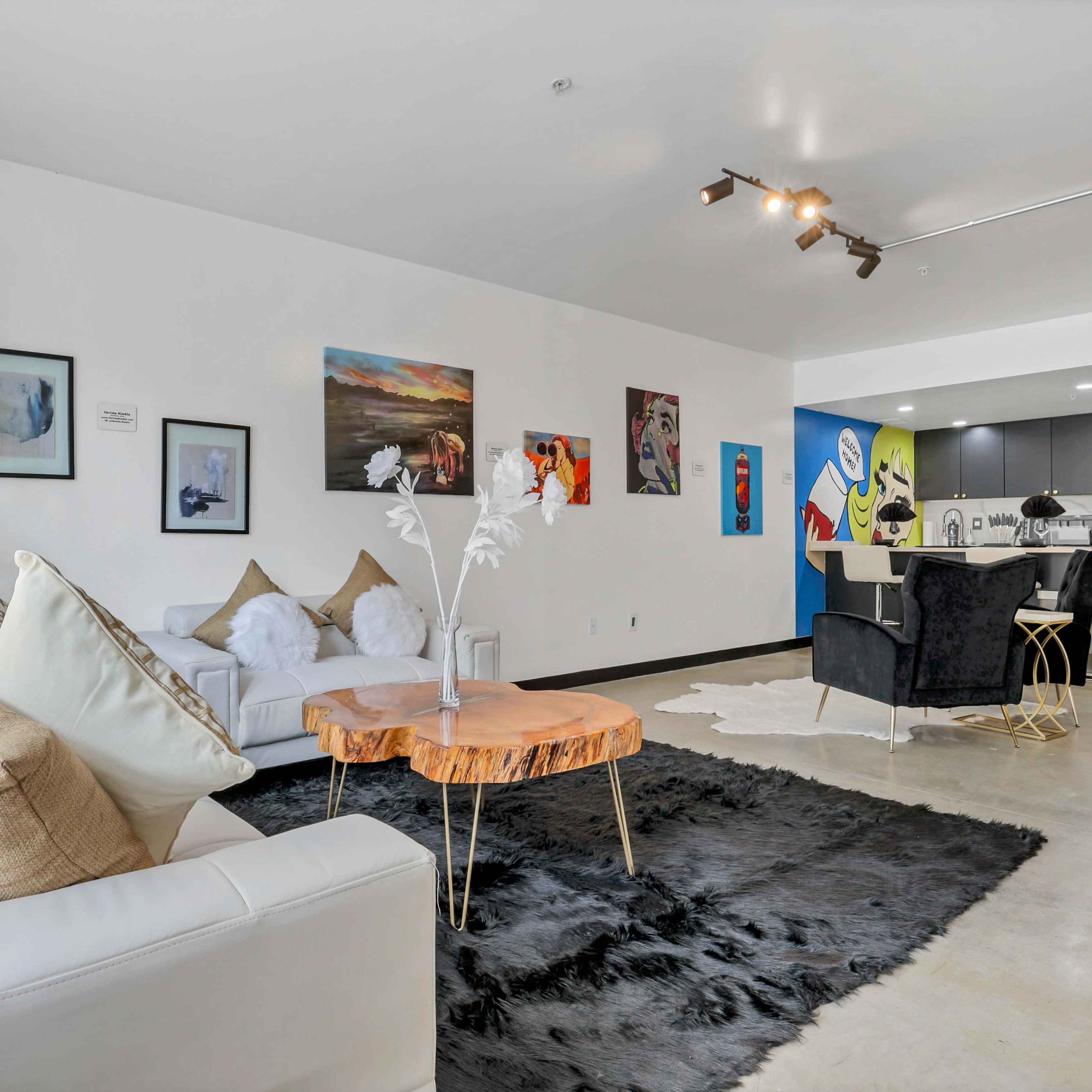 A modern living area featuring a white sofa, a coffee table with a natural wood finish, a black area rug, and colorful artwork on the walls, with a kitchen visible in the background.