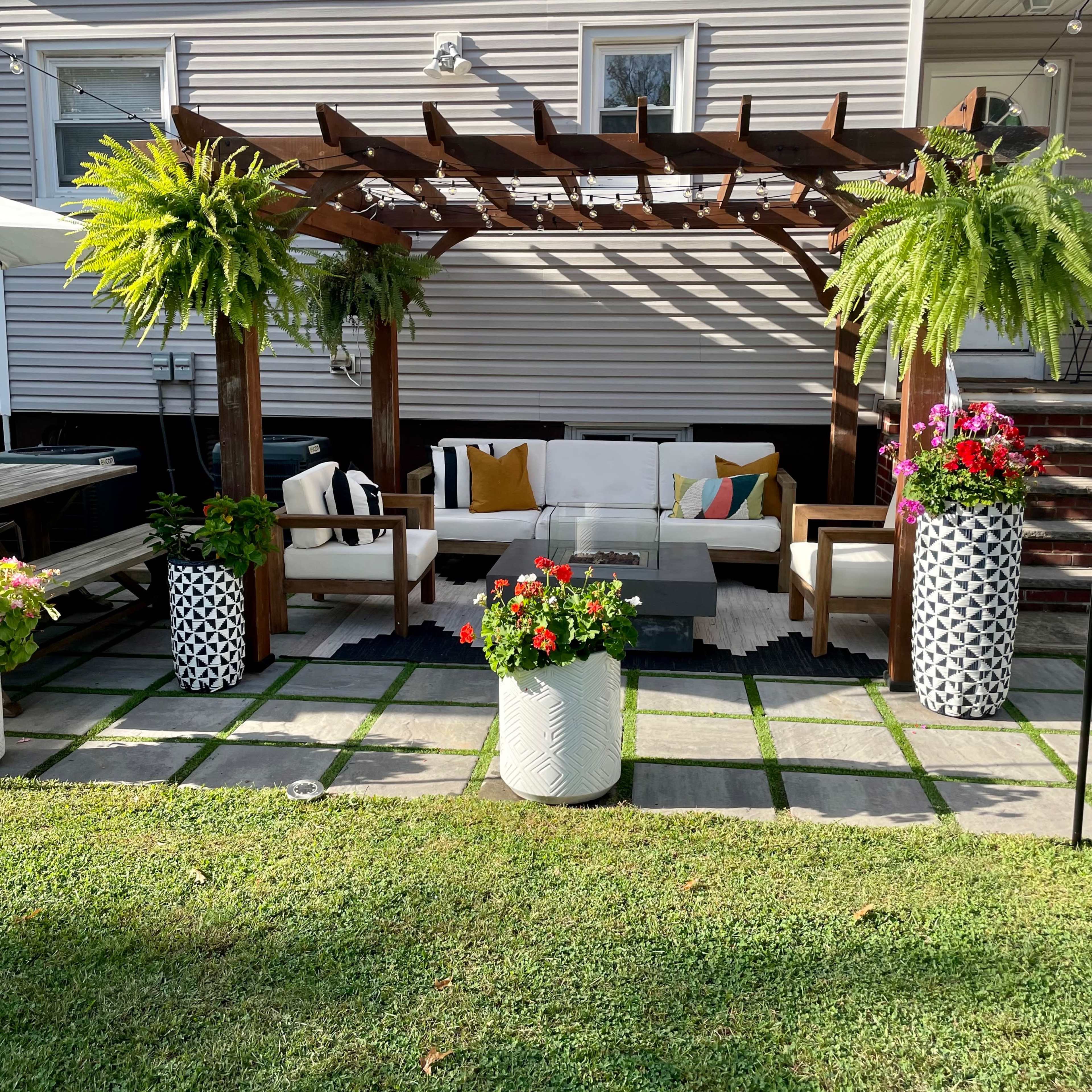 A shaded outdoor seating area with a wooden pergola draped in ferns, featuring a white couch and colorful cushions, surrounded by potted floral arrangements on a grass lawn.