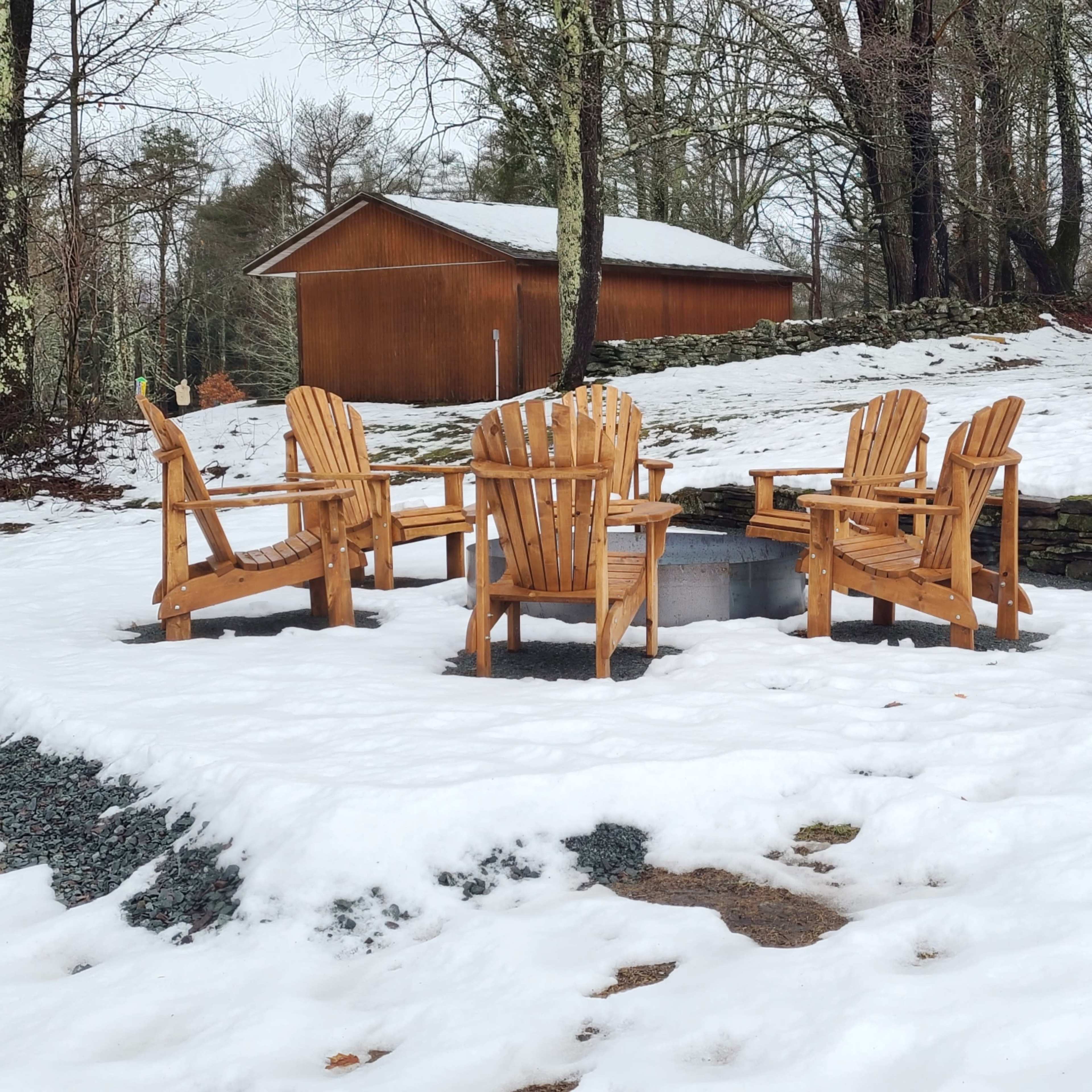 Four wooden chairs arranged in a circular formation around a fire pit, surrounded by a snowy landscape and a wooden shed in the background.