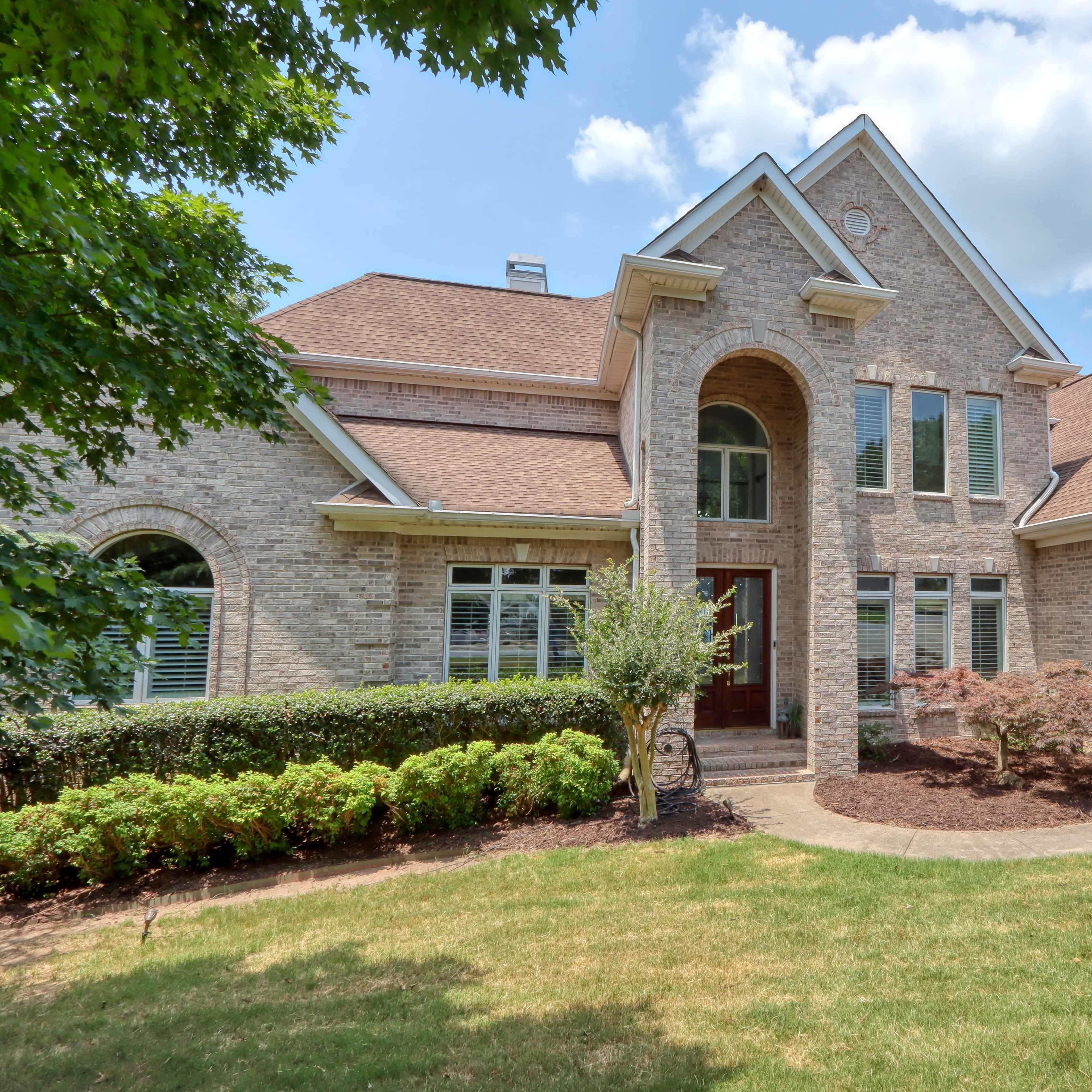 A large brick house with a peaked roof, multiple windows, and a two-car garage is surrounded by neatly trimmed shrubs and a grassy lawn.