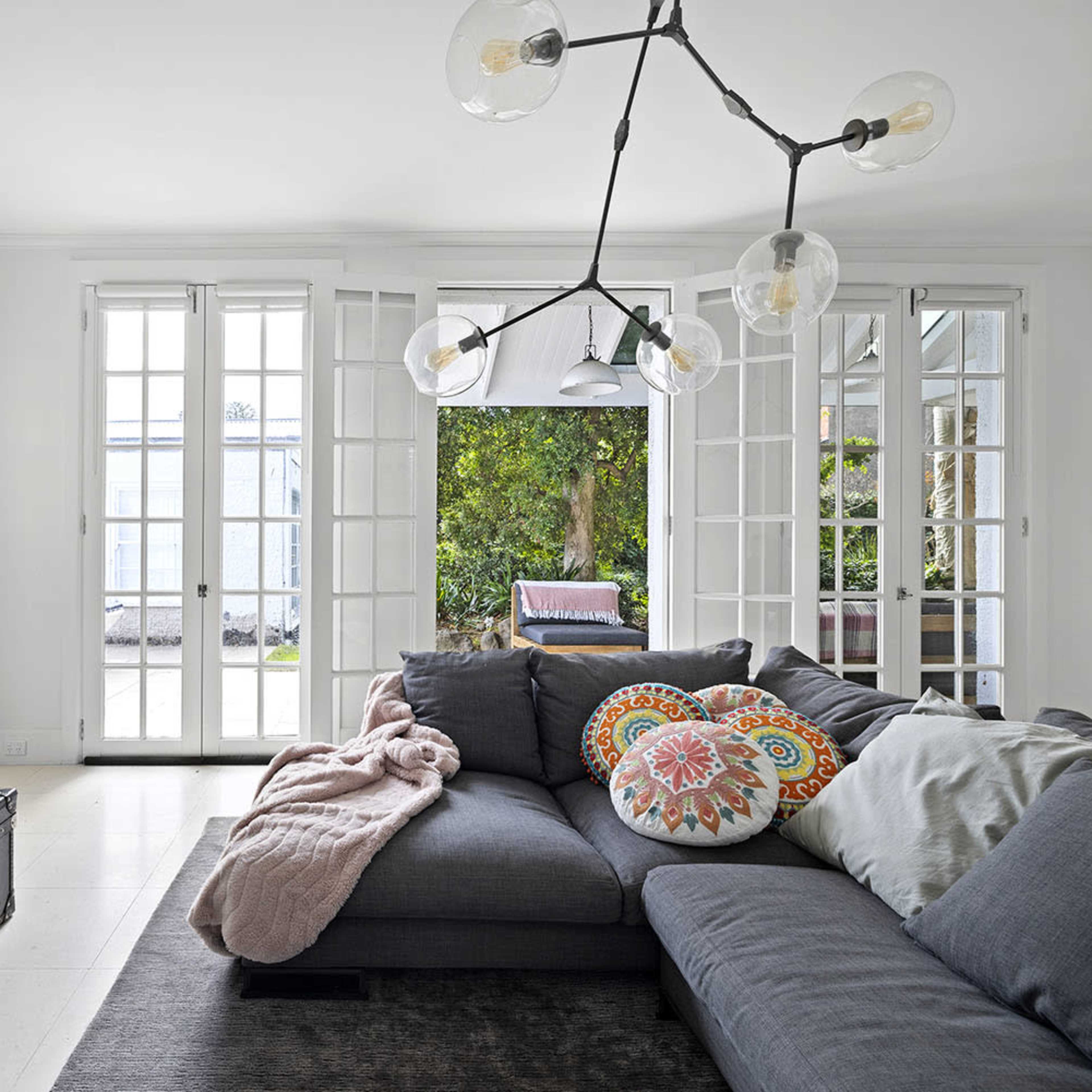 A modern living room features a gray sectional sofa with decorative cushions, a vintage suitcase-style coffee table, and large windows opening to a garden area.