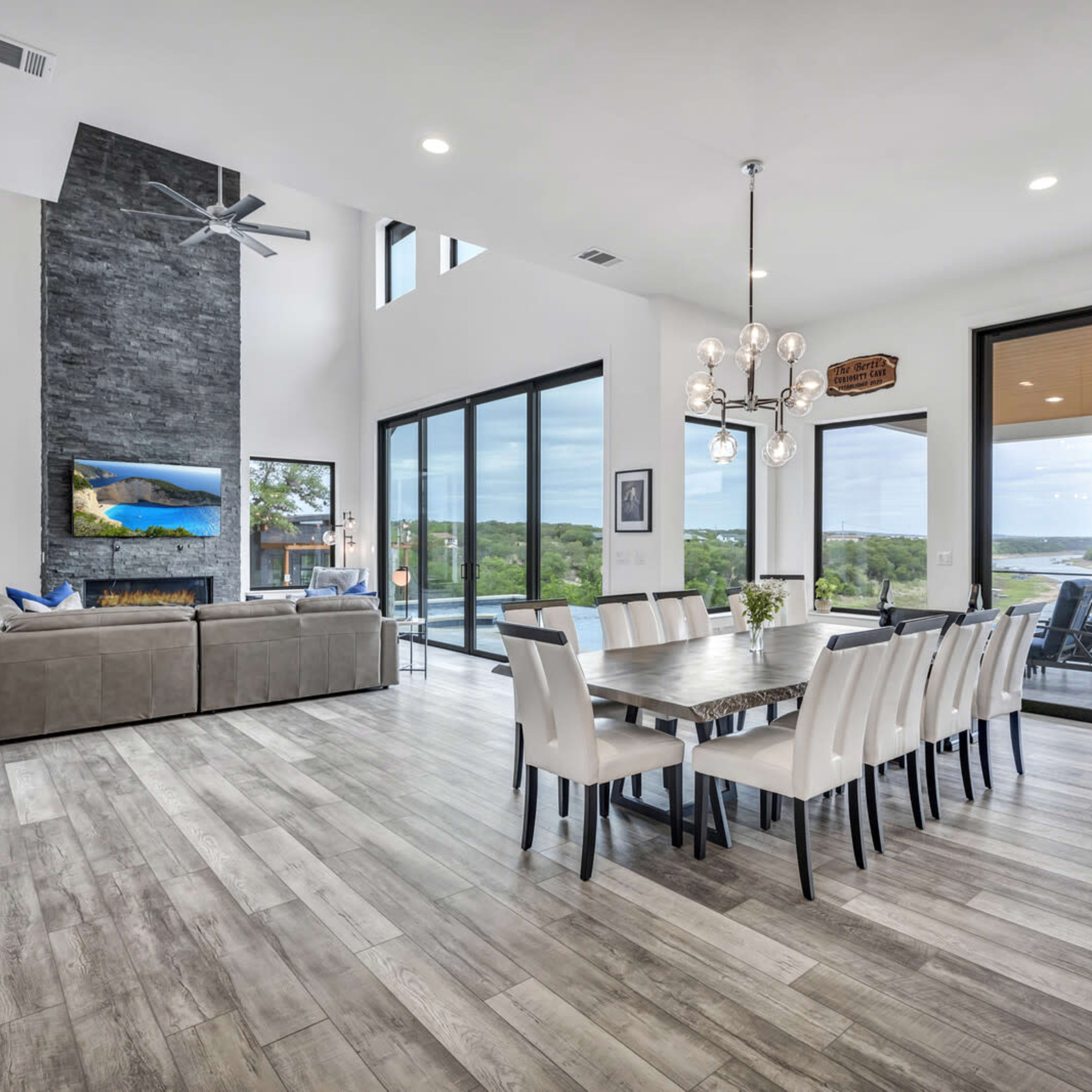A modern, open-concept living and dining area with large windows overlooking a scenic landscape, featuring a long dining table and a sectional sofa.