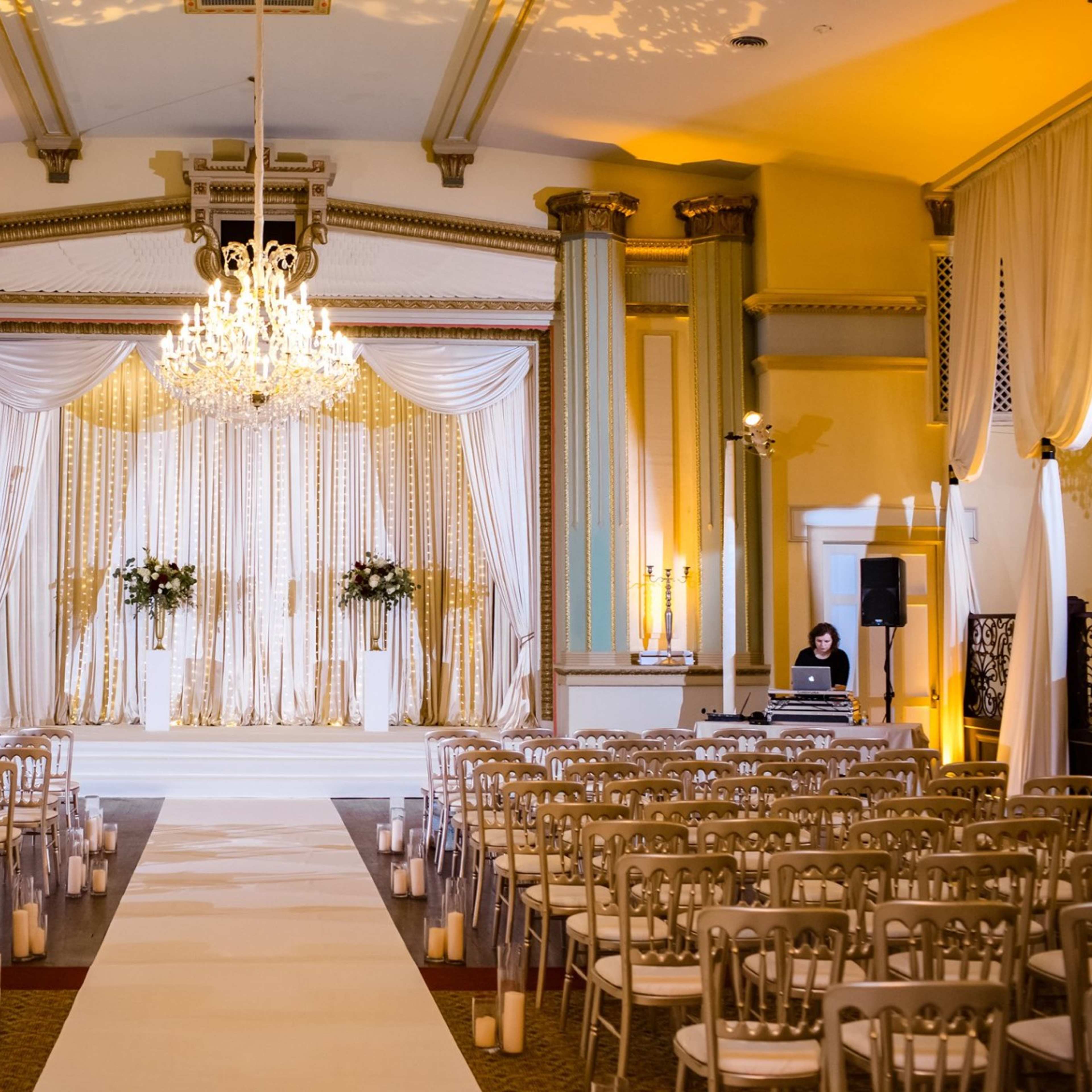 A spacious event hall is set up for a wedding ceremony, featuring rows of chairs facing a decorated altar with a chandelier above.