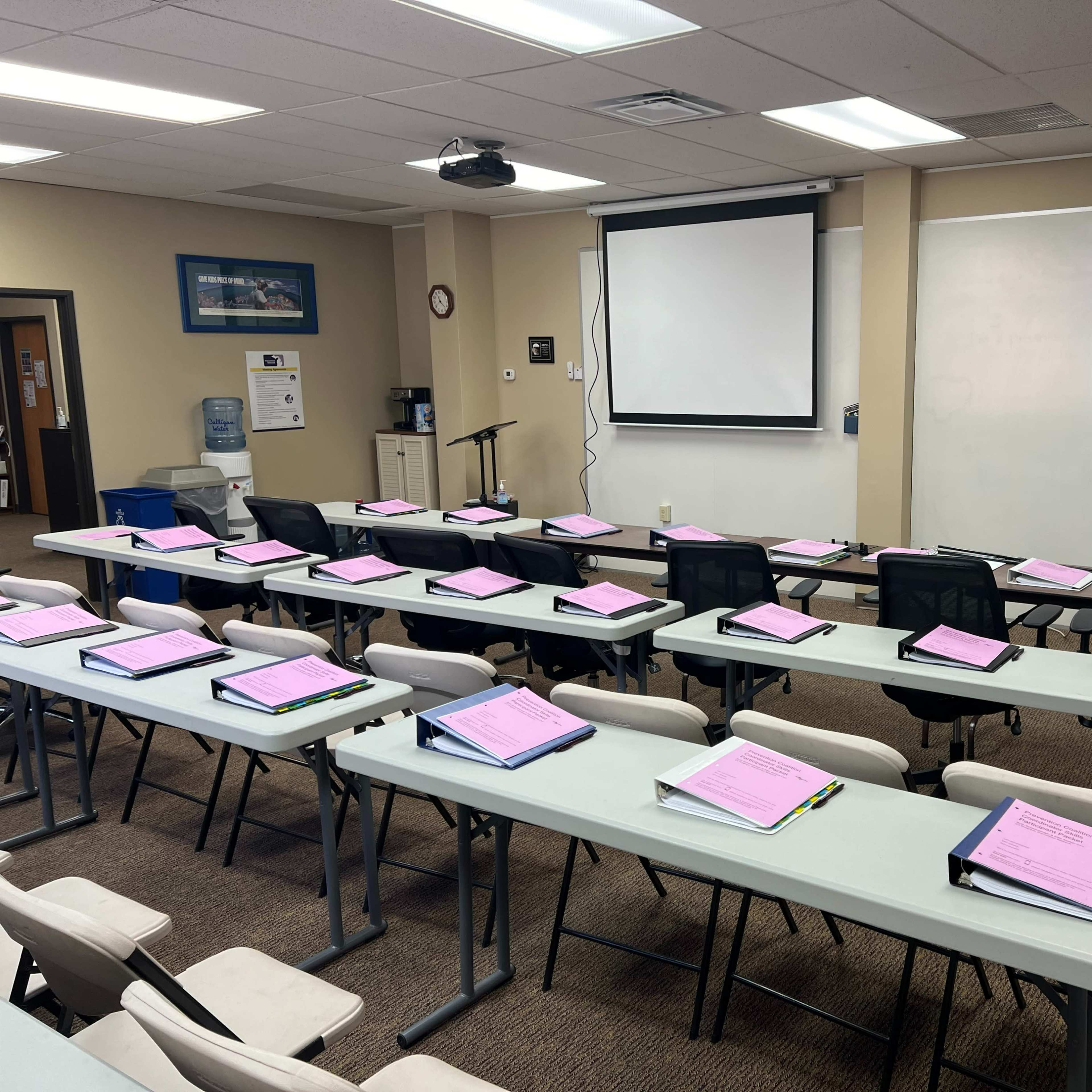A classroom is set up with rows of tables and chairs, each holding a pink folder, and a projector screen is displayed at the front.