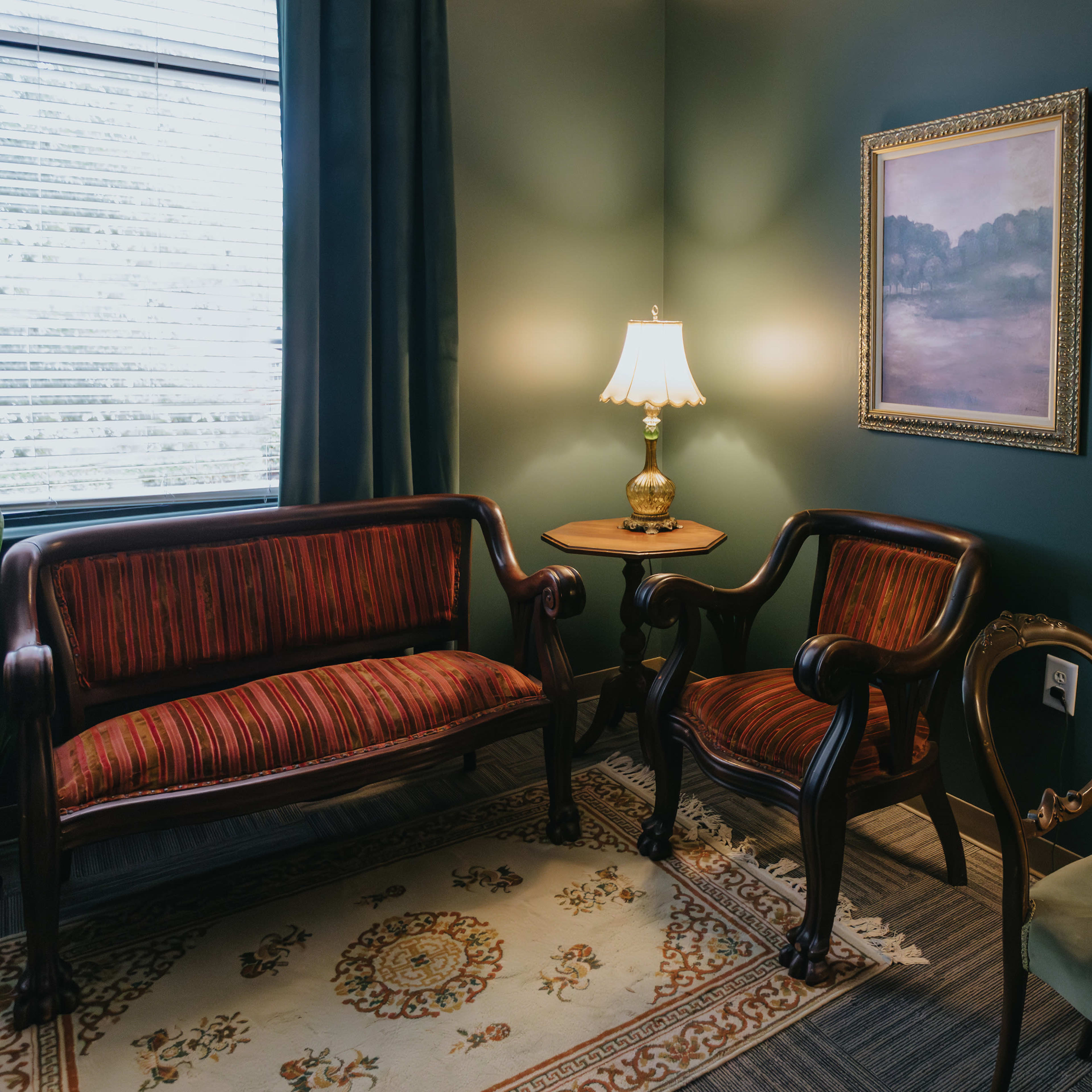 A cozy seating area features two vintage chairs, a small side table with a lamp, and wall art in a green room.