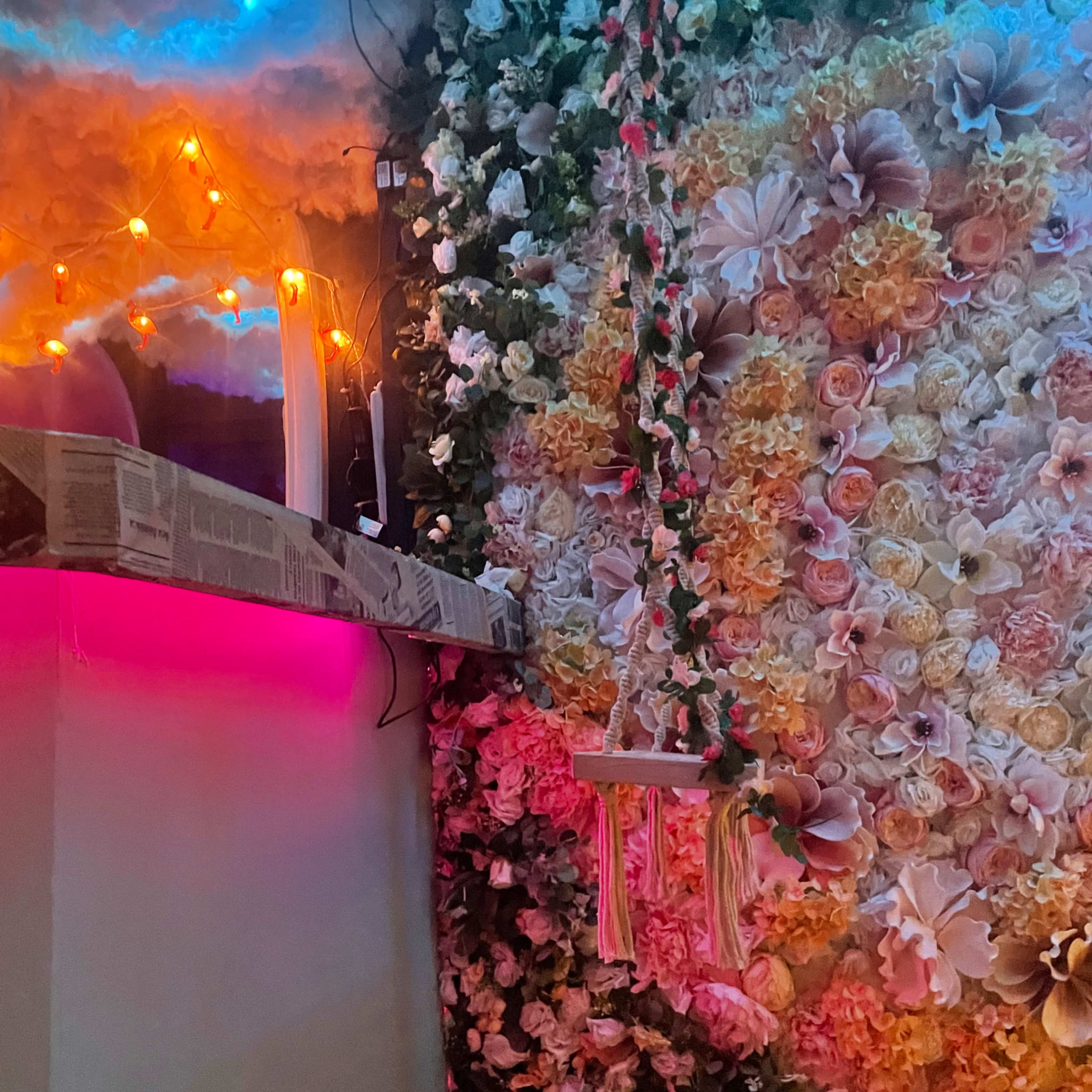 A wall covered in colorful artificial flowers, with a small swing hanging from it and soft lighting illuminating the scene.