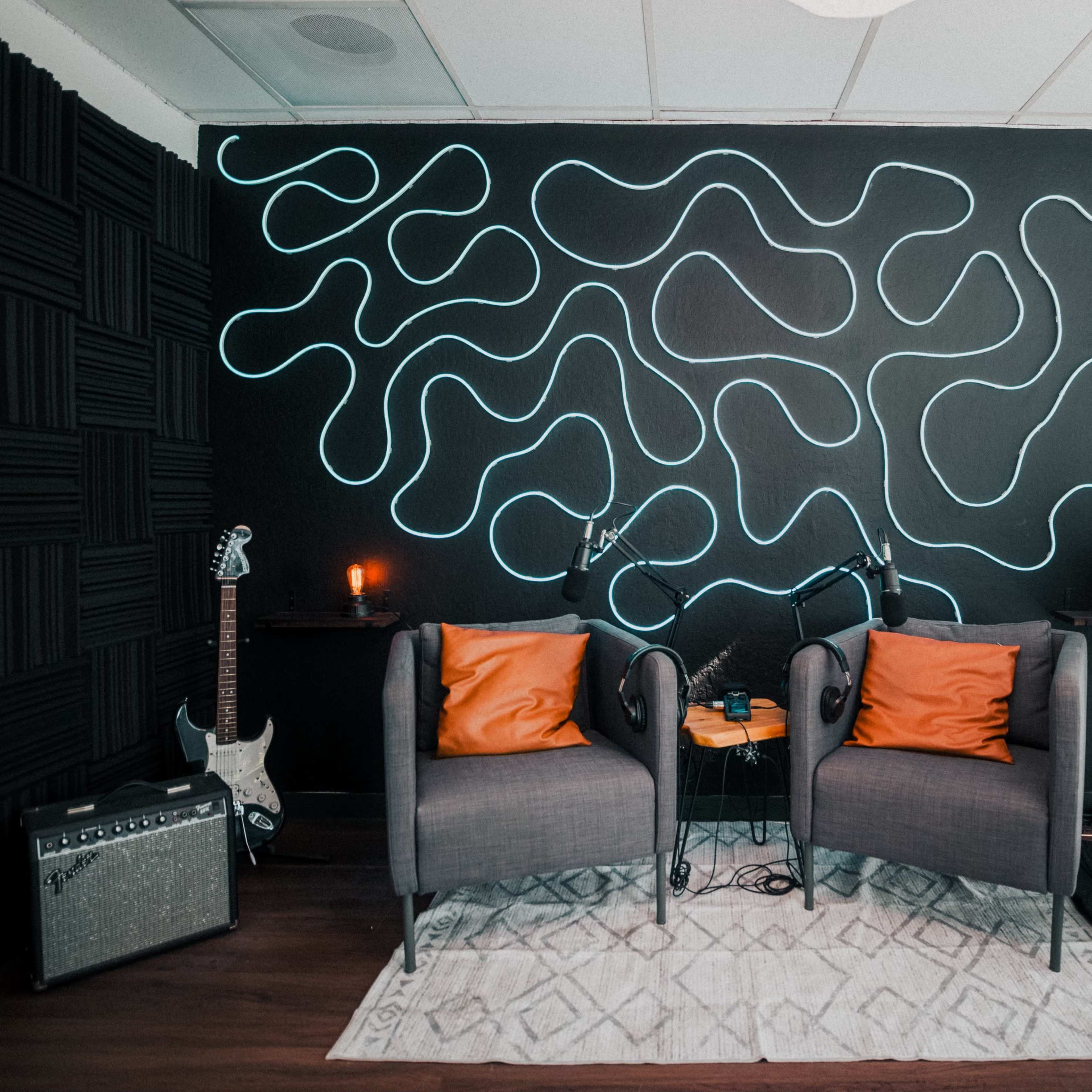 A cozy corner features two gray chairs with orange cushions, a small wooden table, a potted plant, and a wall adorned with wavy neon lights against a dark background.