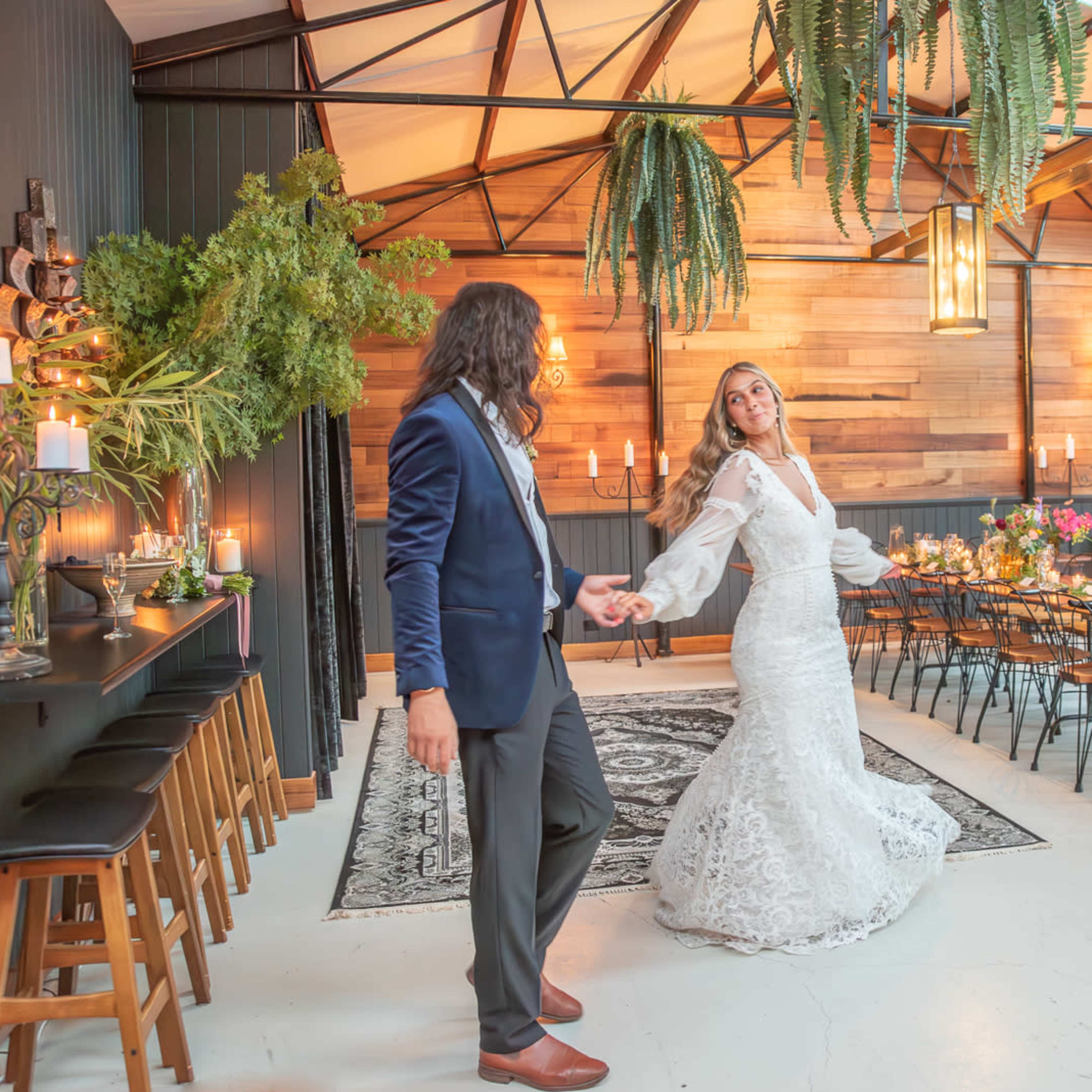 A couple dances in a beautifully decorated indoor venue with wooden walls, greenery, and a long dining table set for a celebration.