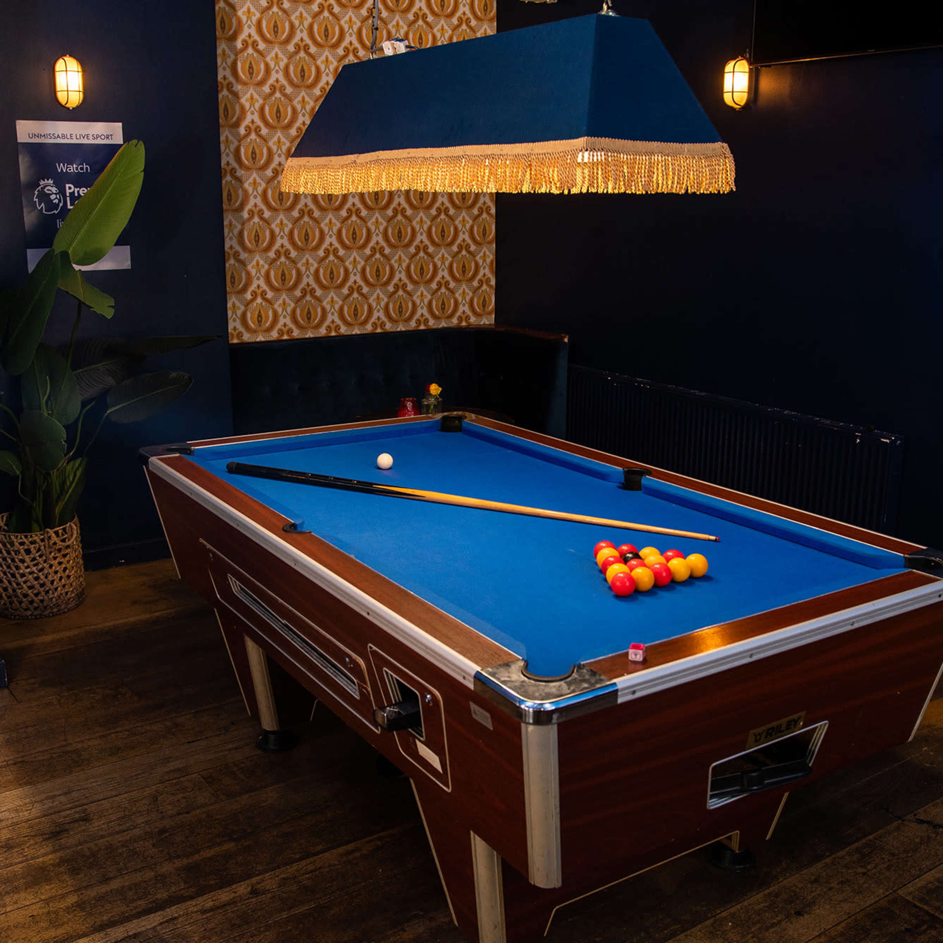 A billiards table set with colorful balls and a cue stick is positioned under a large light fixture in a dimly lit room.
