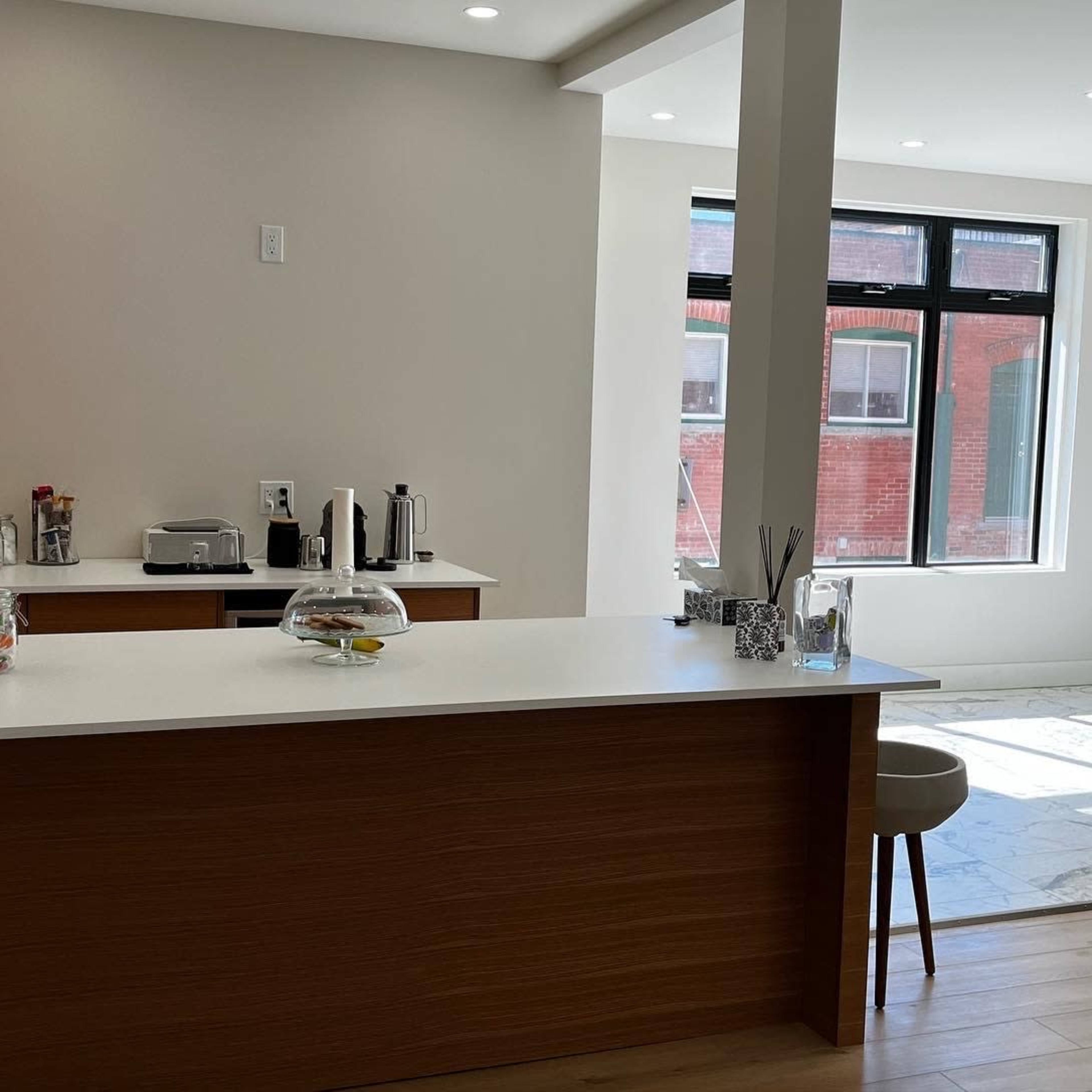 A modern kitchen features a central island with a glass cloche and a seating area, adjacent to a brightly lit dining space with large windows.