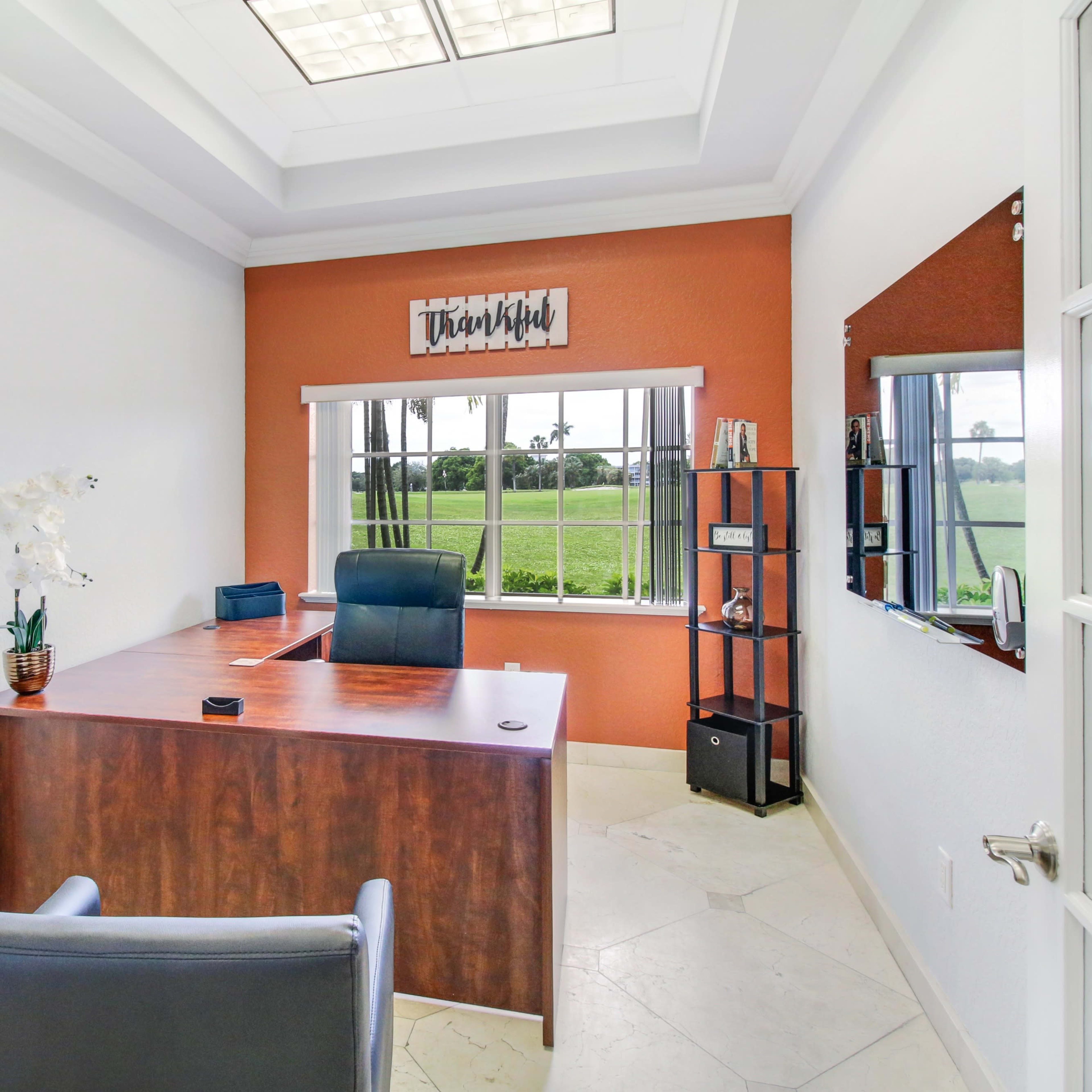 A small office with a wooden desk, two chairs, a bookshelf, and a large window overlooking a green landscape.