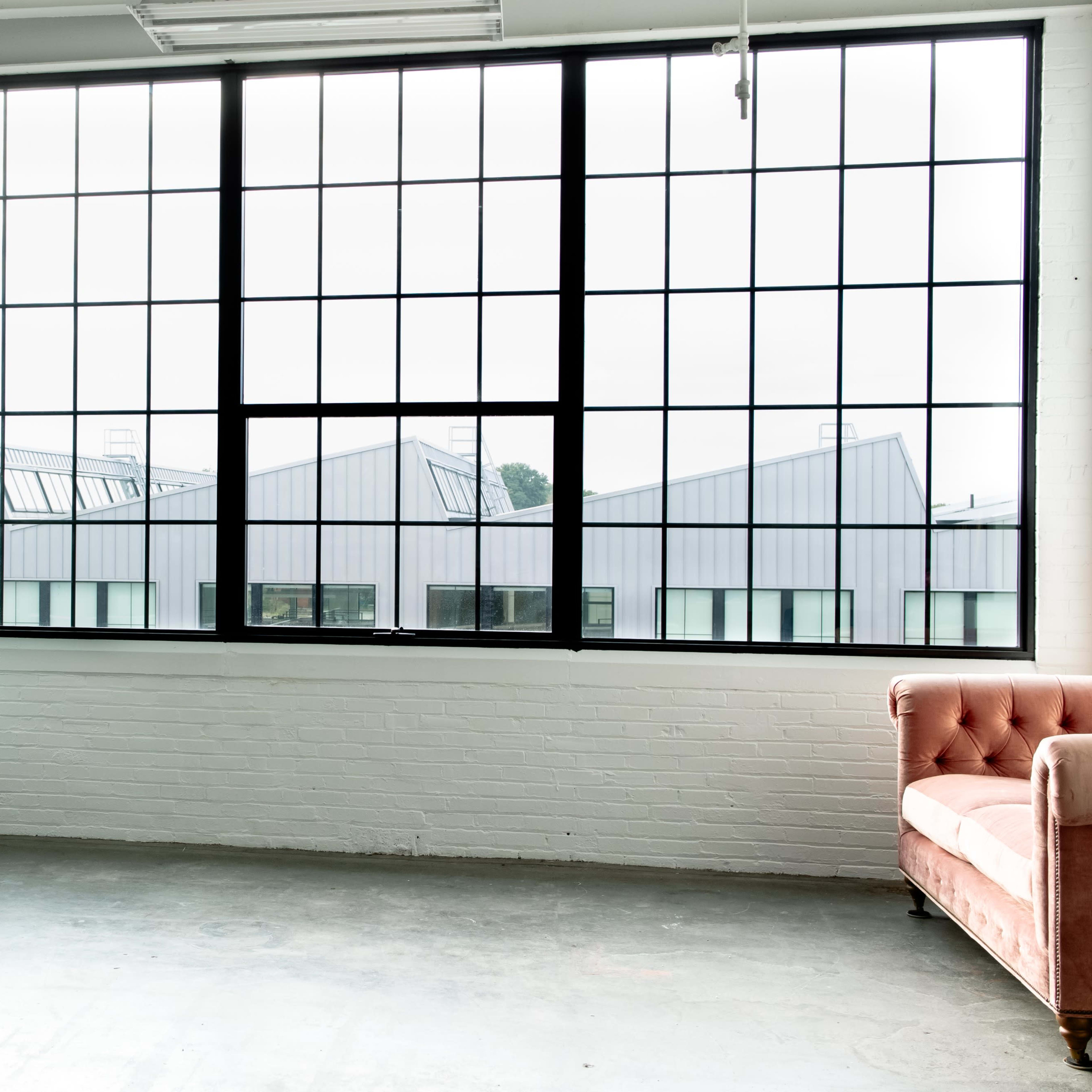 A spacious room with large windows overlooking industrial-style buildings, featuring a green sofa and a pink armchair against the white walls.