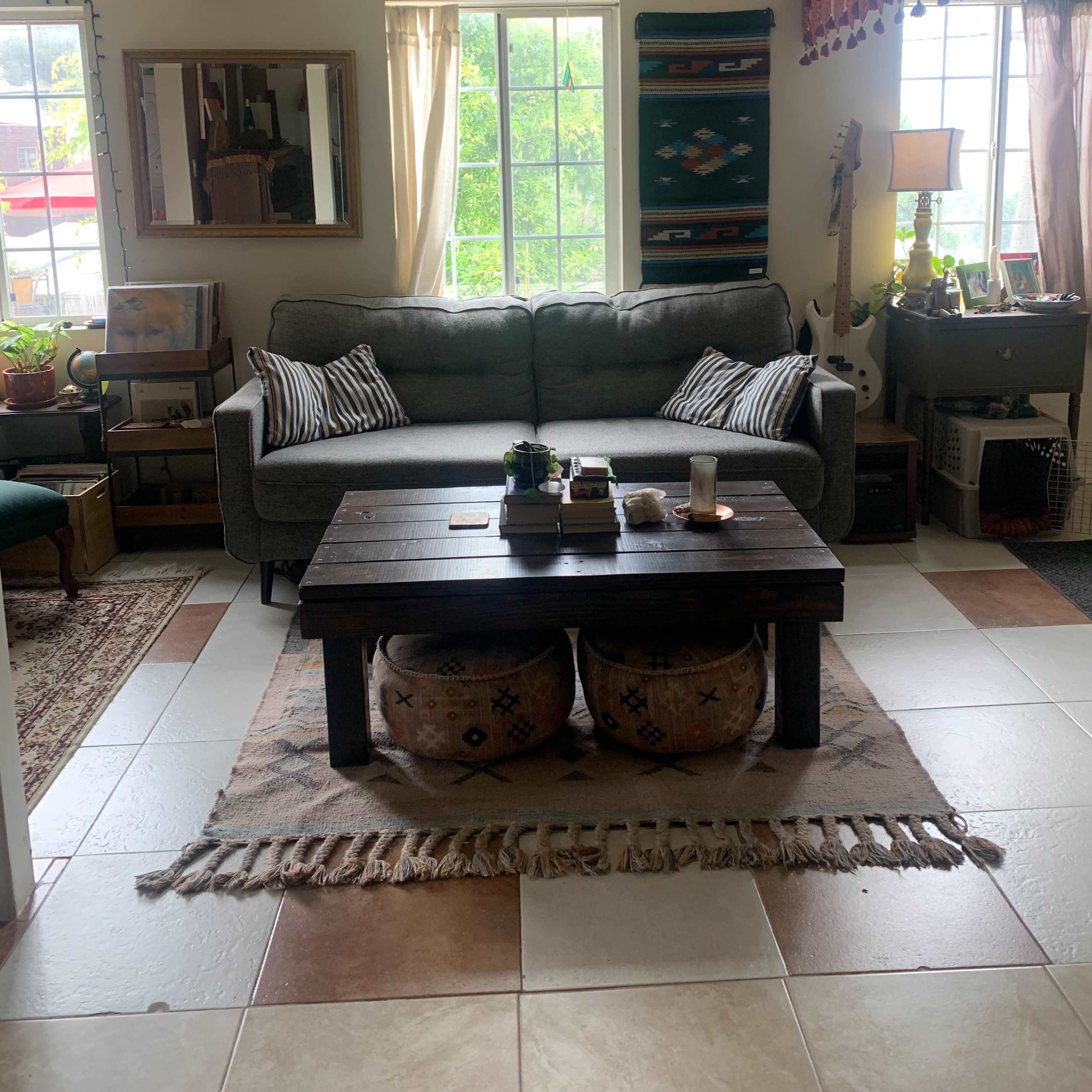 A cozy living room featuring a gray sofa, a wooden coffee table with decorative items, two upholstered stools, and a bright window with plants and artwork around.
