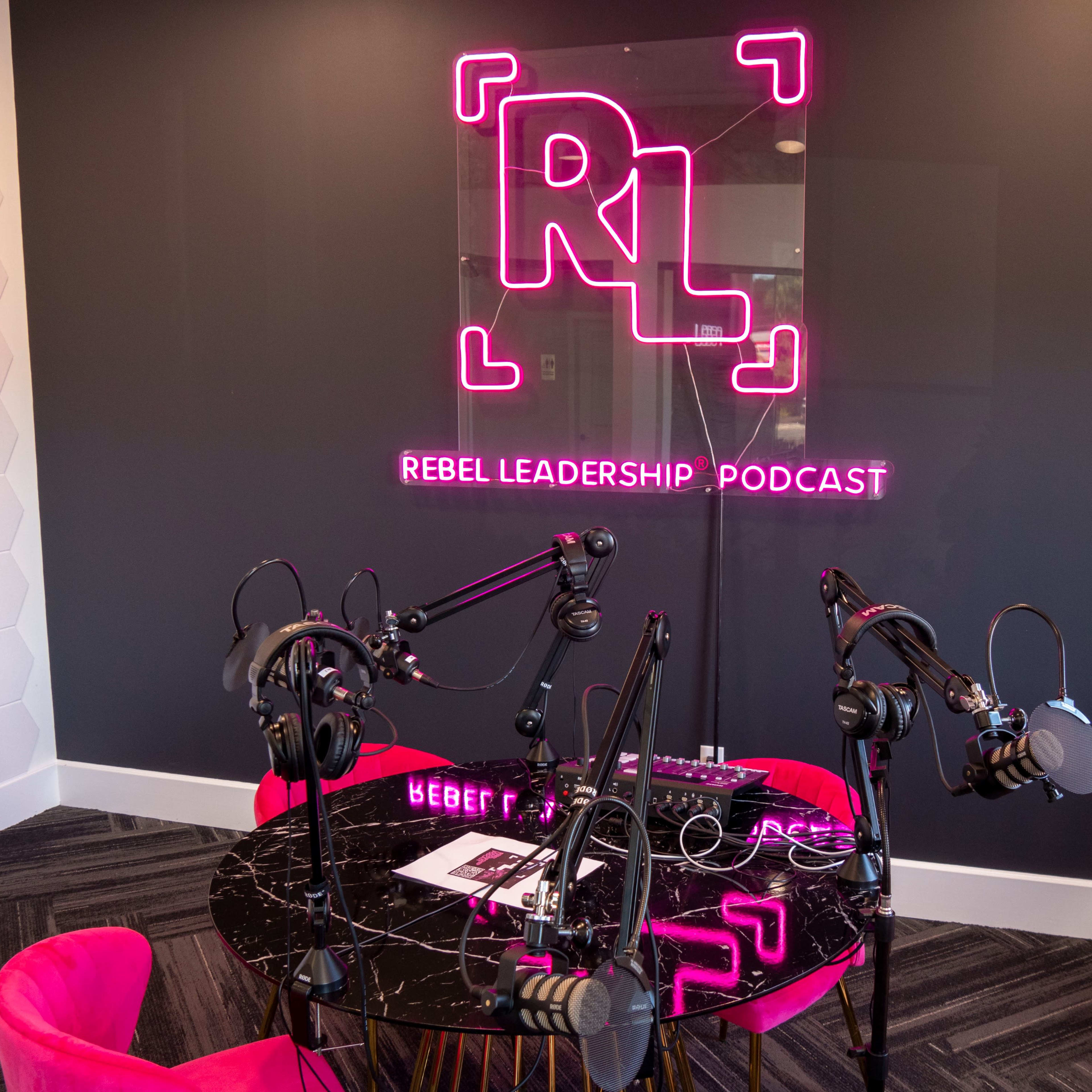 A podcasting studio featuring a black table with multiple microphones and headphones, illuminated by a neon sign that reads "REBEL LEADERSHIP PODCAST."