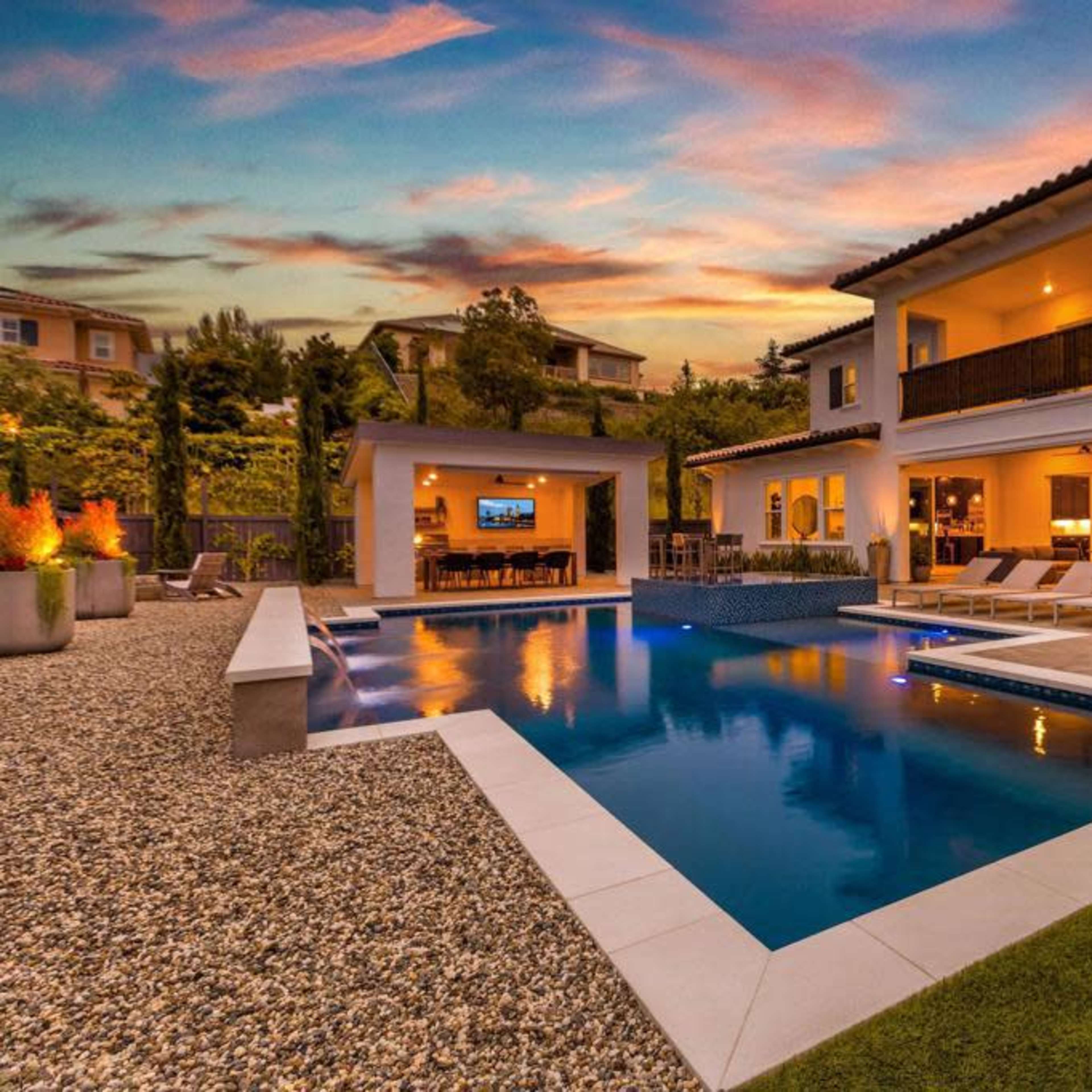 A modern backyard with a swimming pool, lounge chairs, and a spacious home illuminated at sunset.