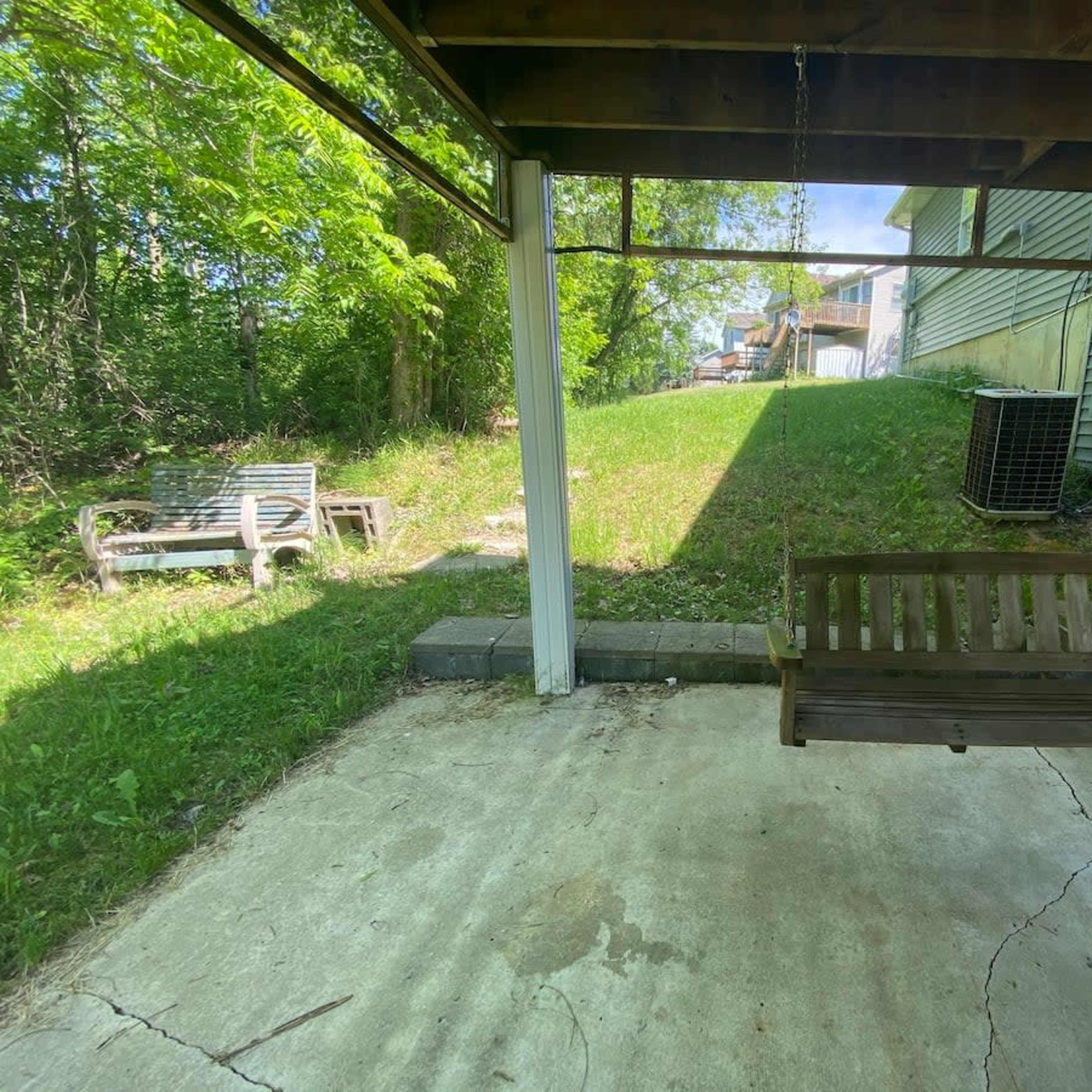 A wooden swing suspended under a shaded porch beside a grassy area, with a lawn chair and a house in the background.