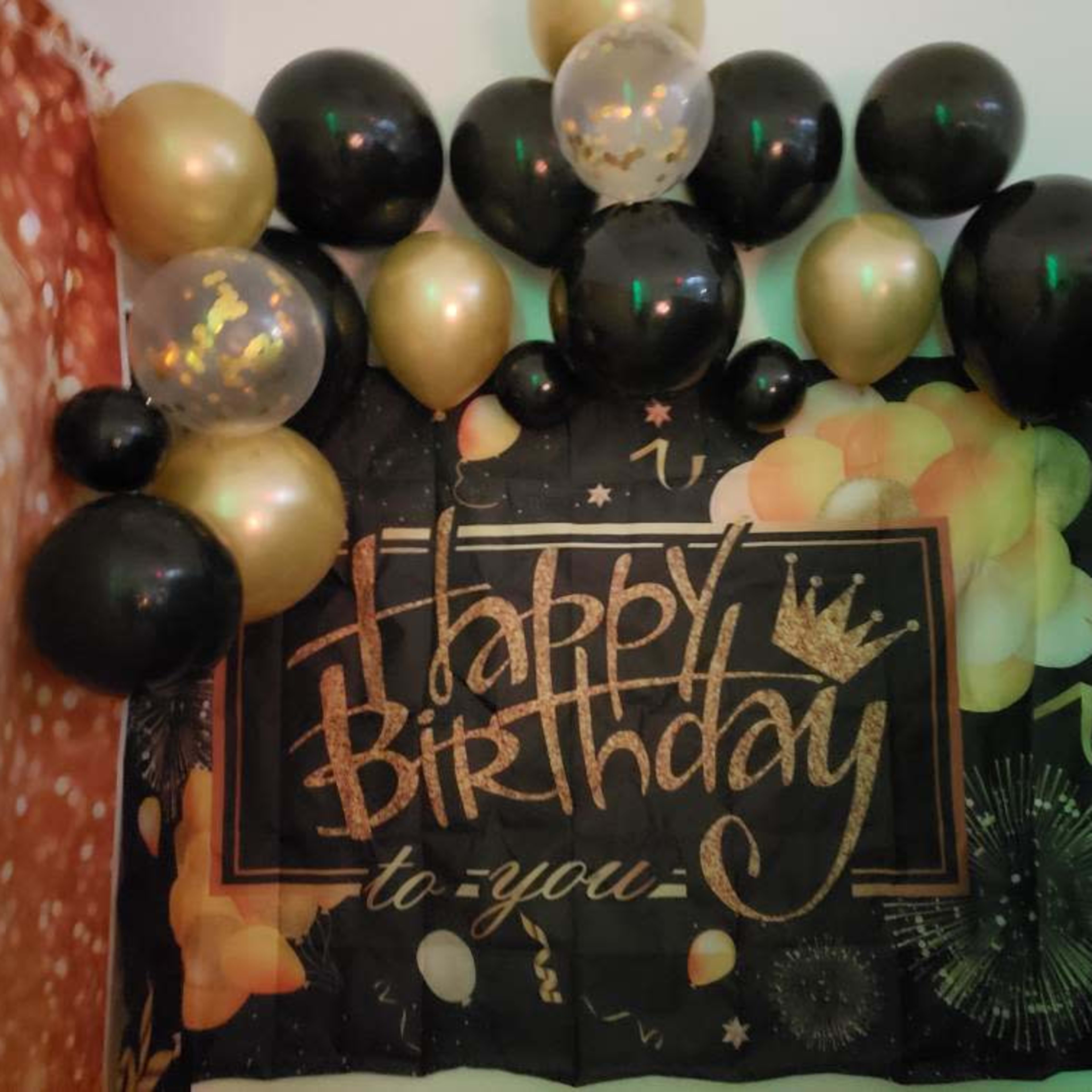 A birthday decoration featuring a large banner reading "Happy Birthday to You" surrounded by black, gold, and transparent balloons.