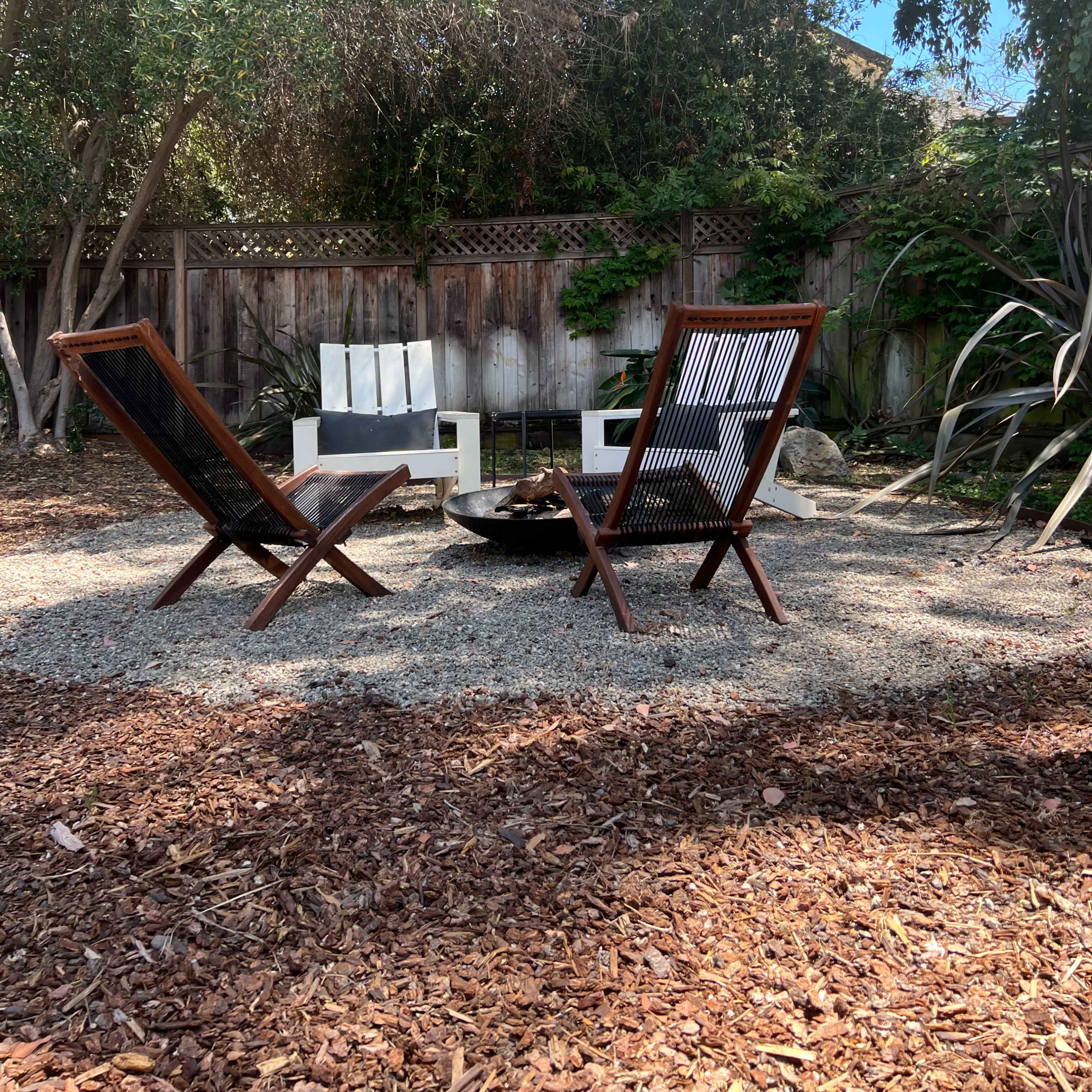 Two wooden chairs face a circular fire pit in a gravelly backyard surrounded by trees and a wooden fence.