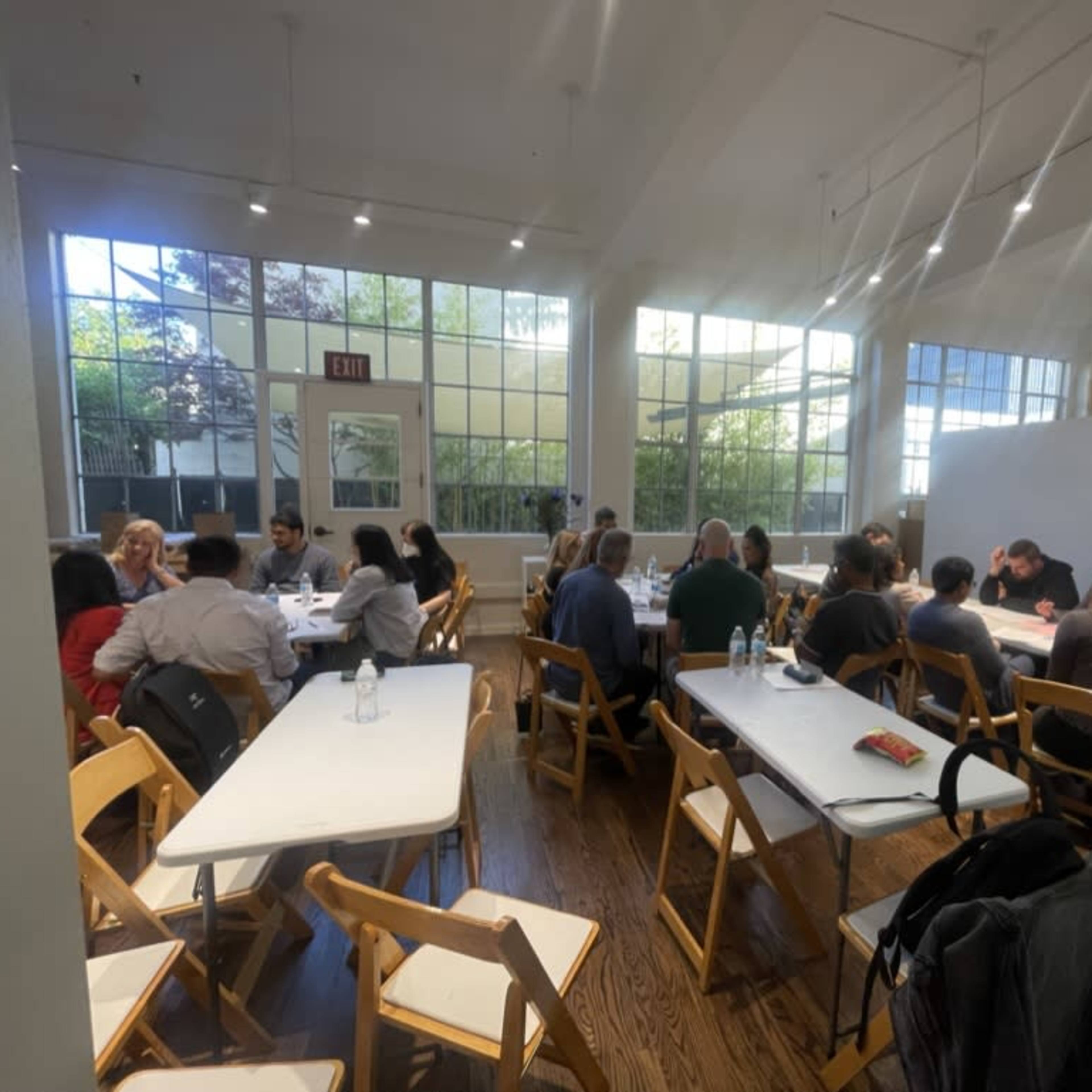 A group of people is seated at tables in a well-lit room with large windows, engaged in discussion.