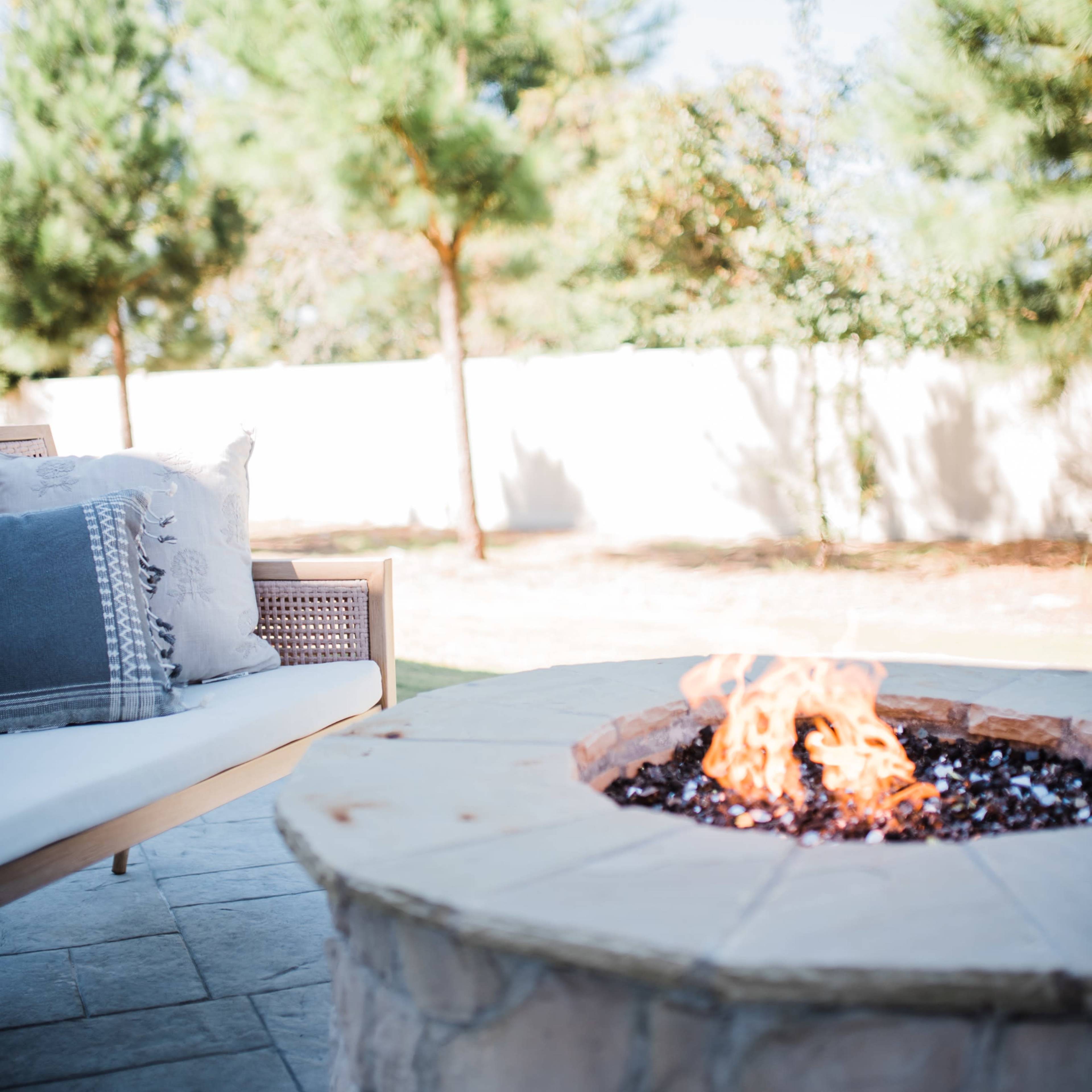 A stone fire pit with flames burns in a backyard surrounded by trees and a white fence.