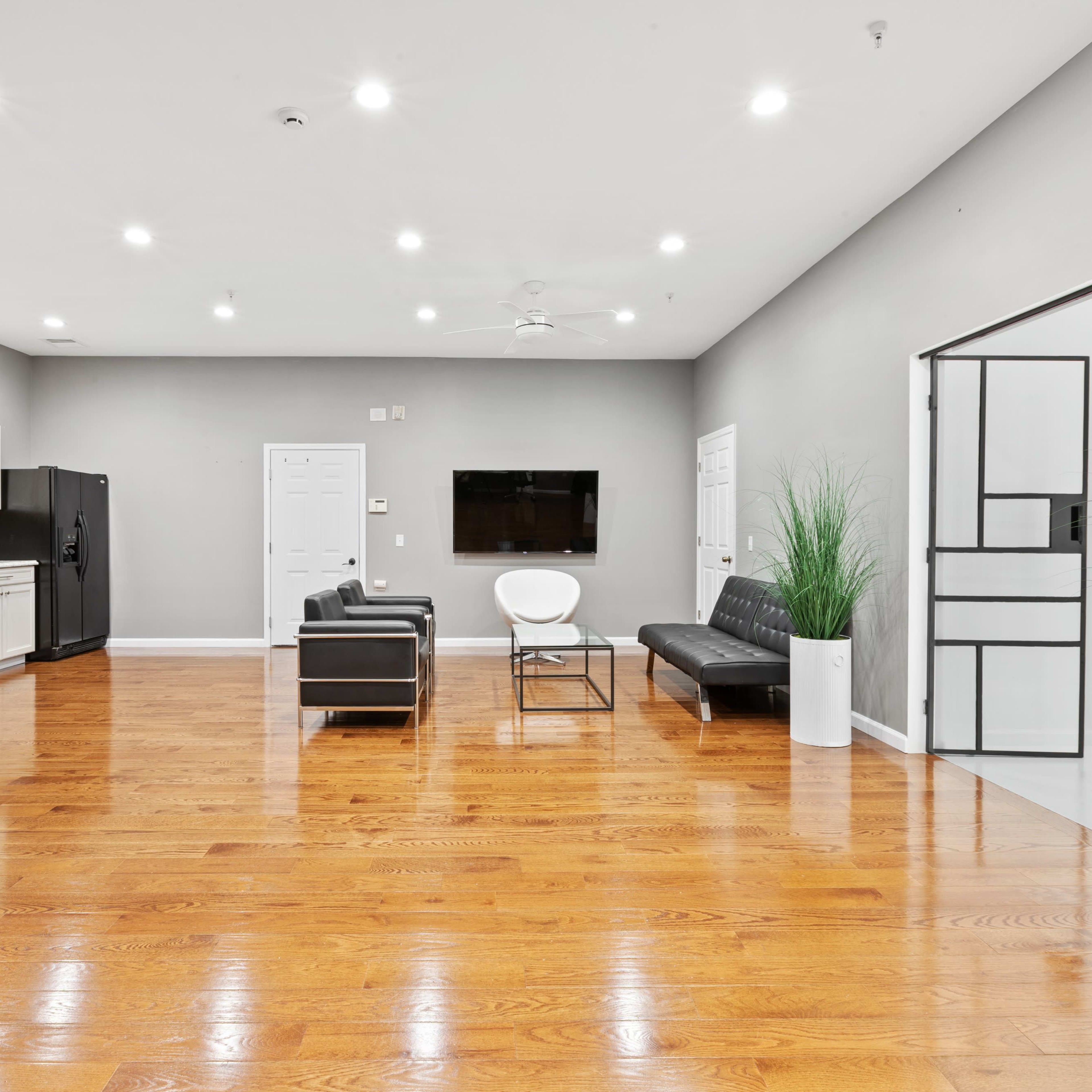 A spacious, modern room features hardwood flooring, a seating area with two black chairs, a white chair, and a wall-mounted television, alongside kitchen cabinets and appliances.