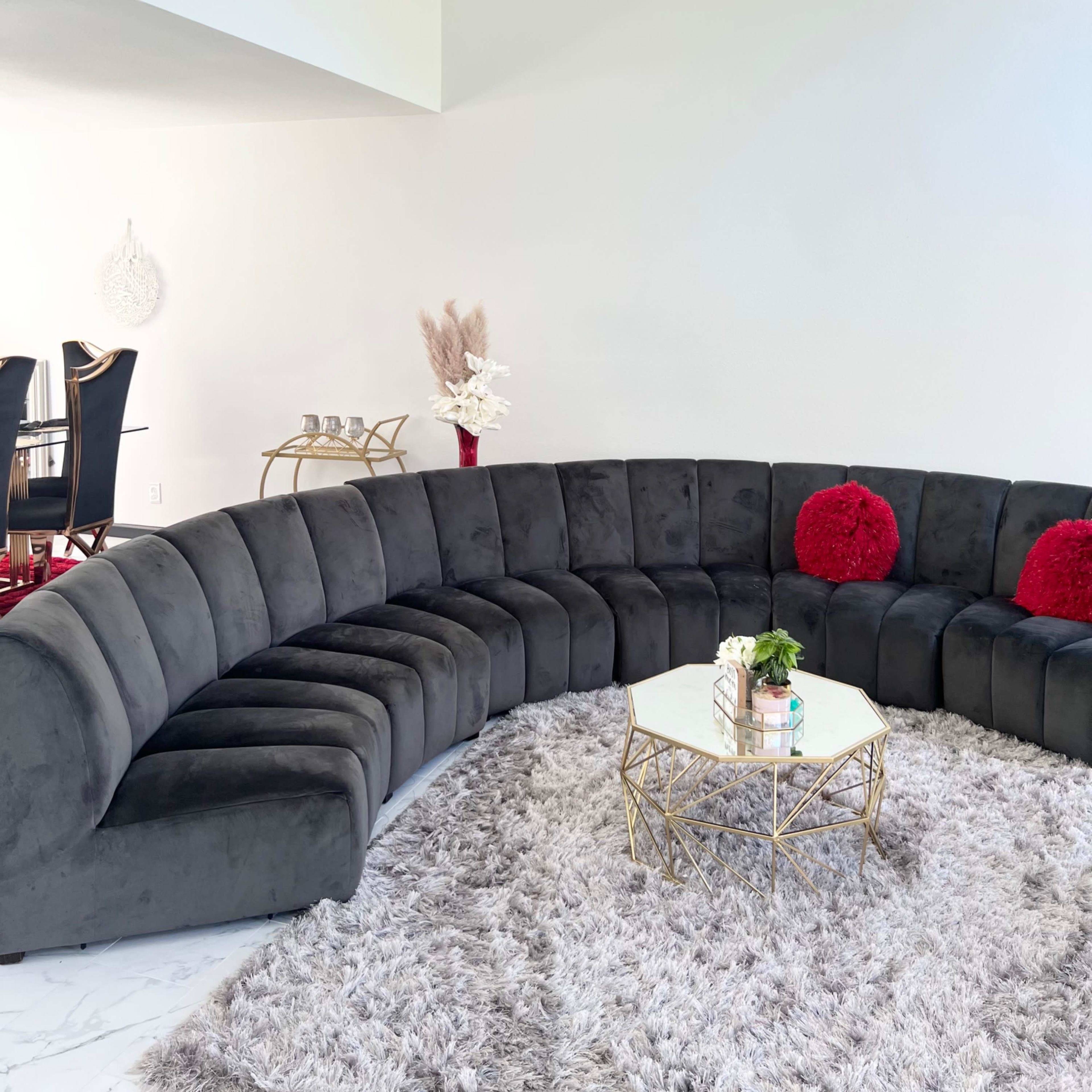 A curved gray sectional sofa arranged around a round coffee table on a textured rug in a bright room.