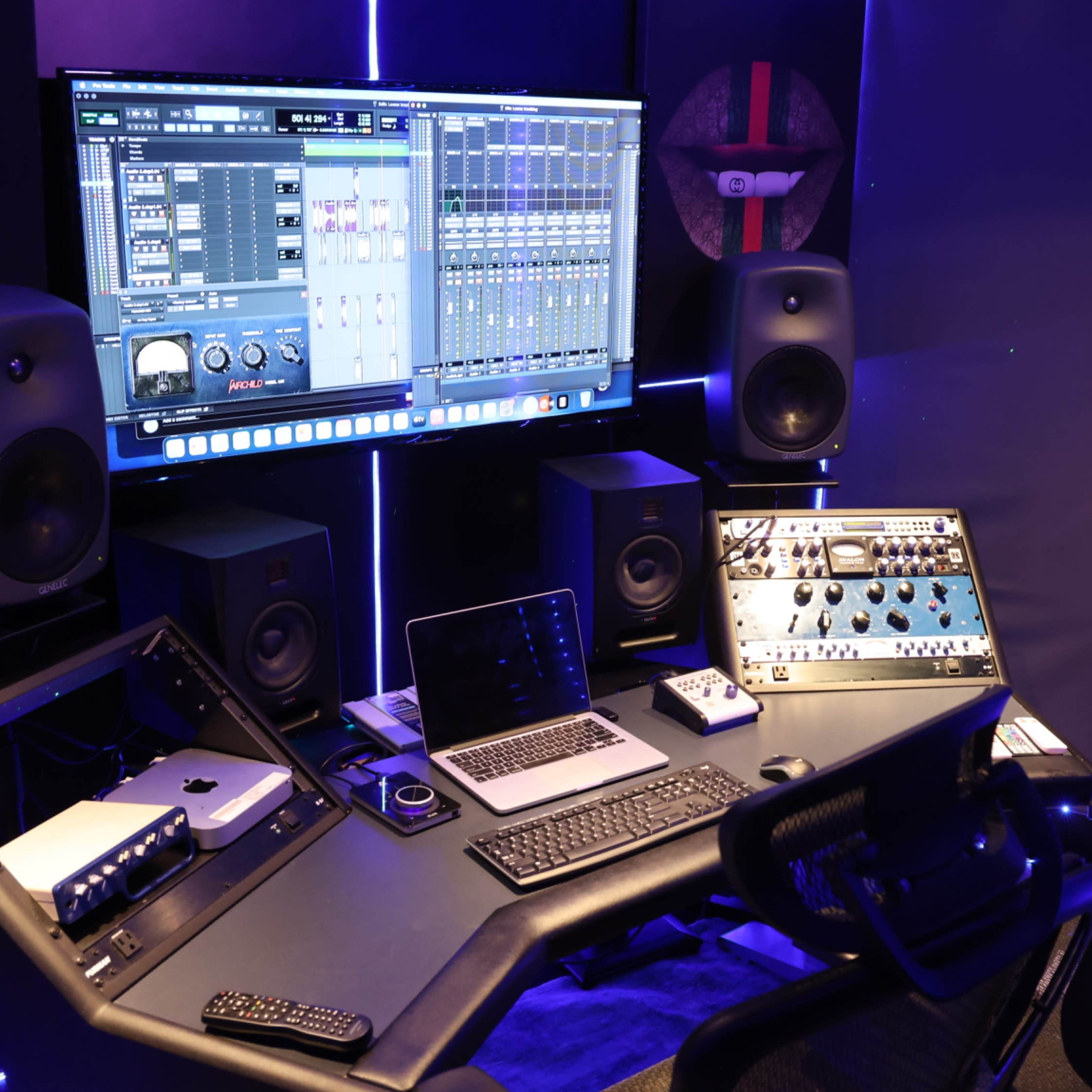 A music production studio features a large monitor displaying audio software, speakers, and a desk equipped with various electronic devices.