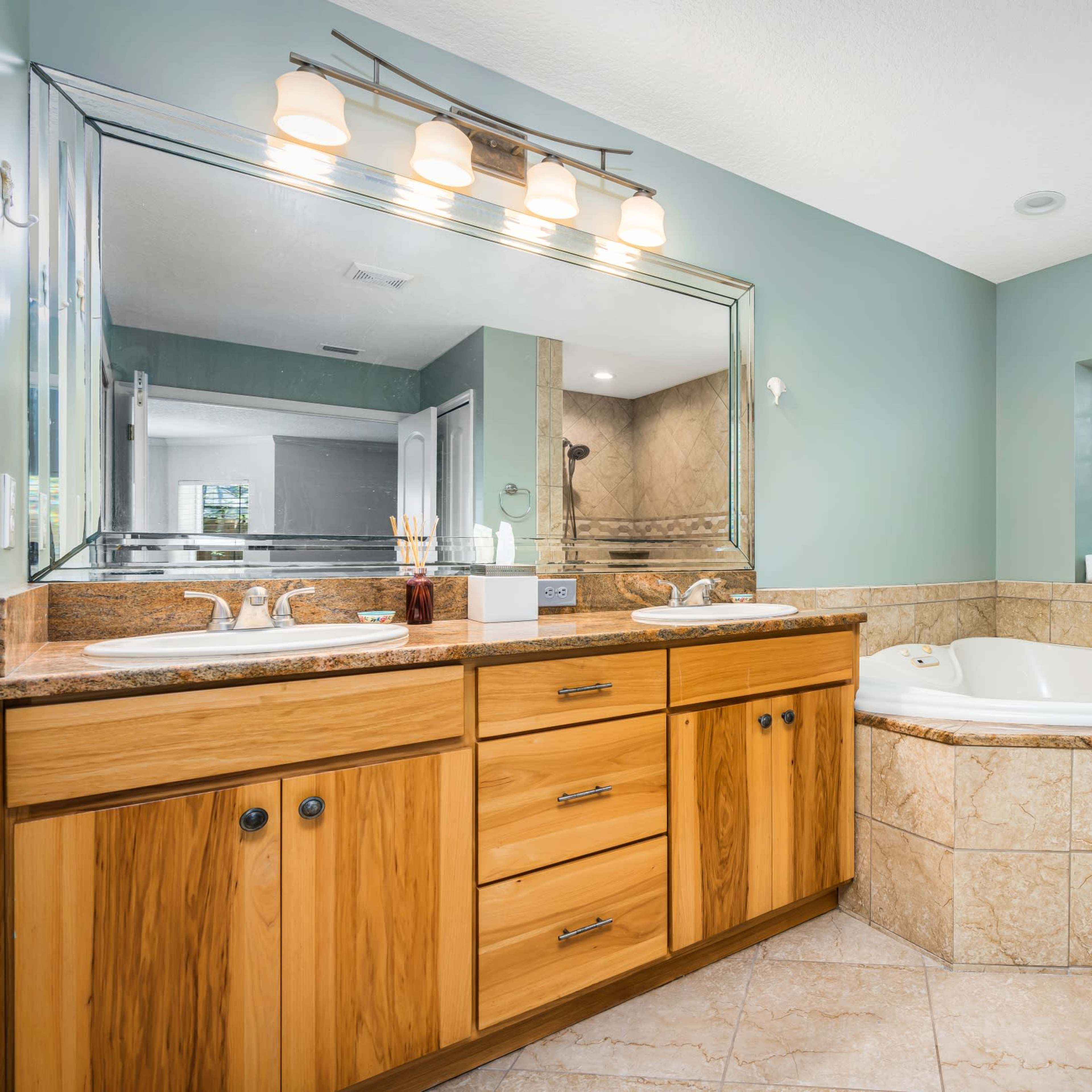 A spacious bathroom featuring dual sinks, a large mirror, a corner jacuzzi tub, and light-colored tiled walls and floors.
