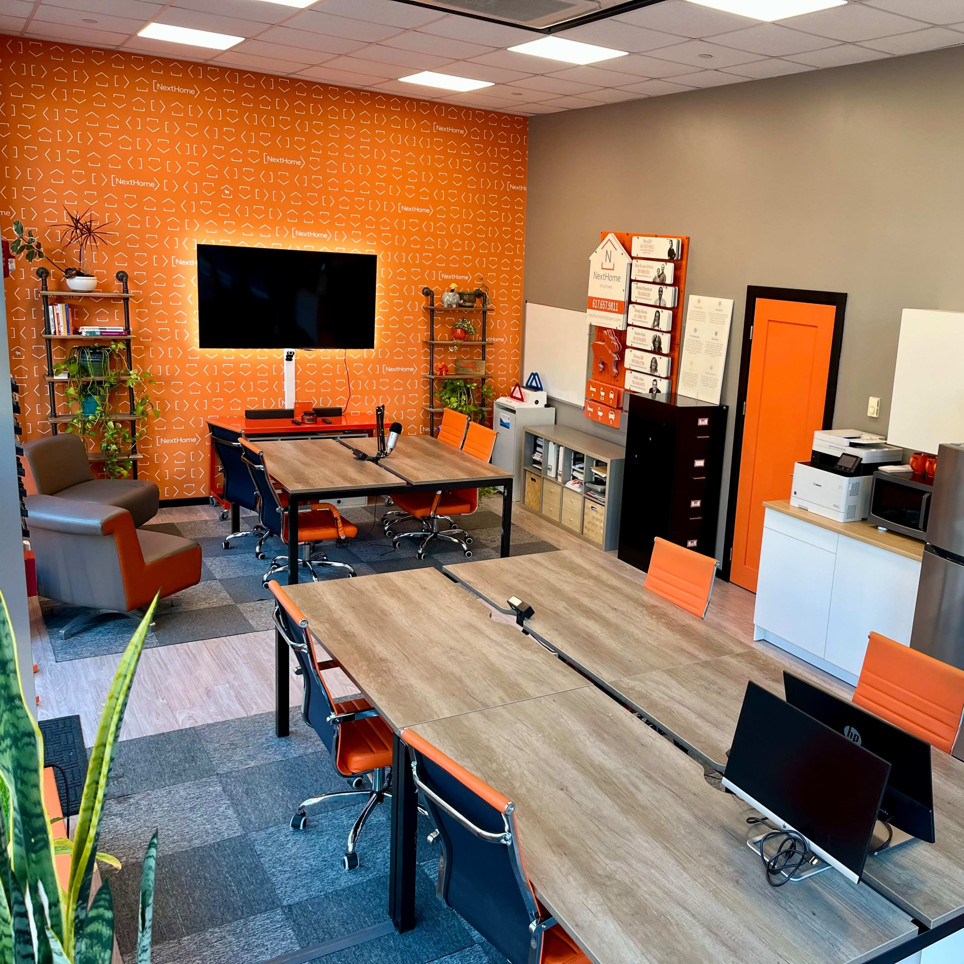 A modern office space featuring several large tables, an orange accent wall, and a kitchen area with a refrigerator and printer, all arranged for collaborative work.