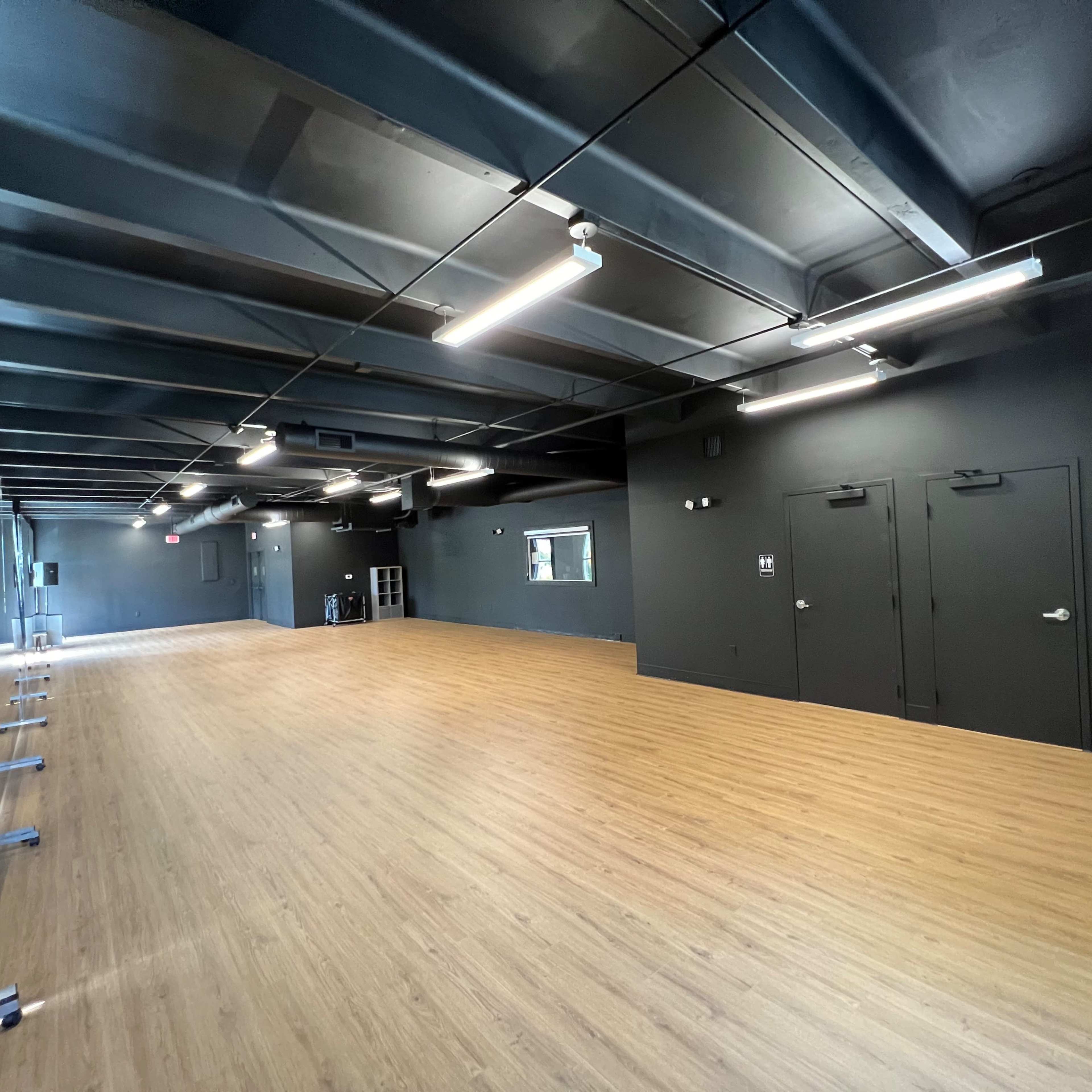 A spacious, empty dance studio featuring a wooden floor, black walls, and a large mirror along one side.