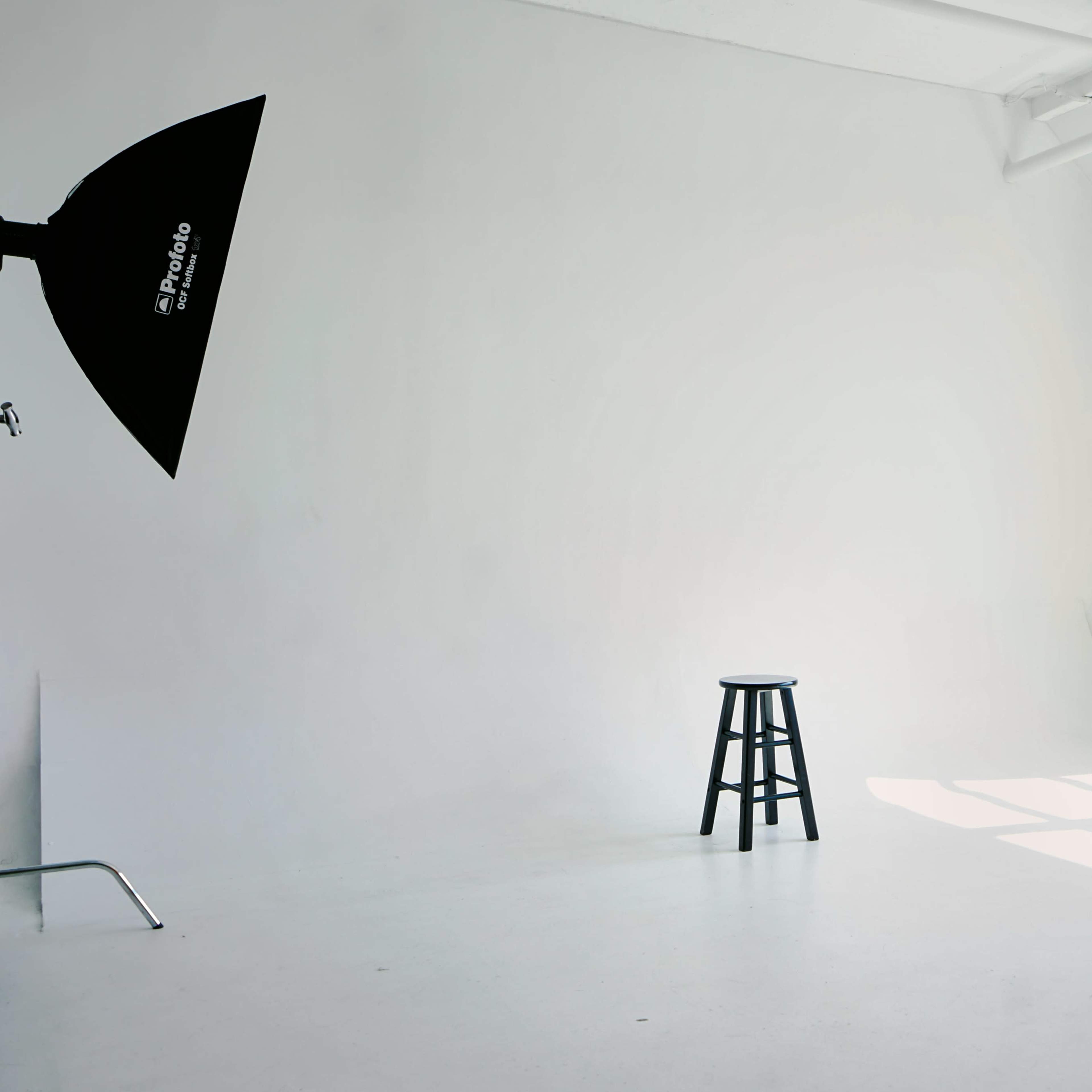 A bright, empty photo studio features a black stool, a light source, and a few boxes, with large windows allowing sunlight to stream in.