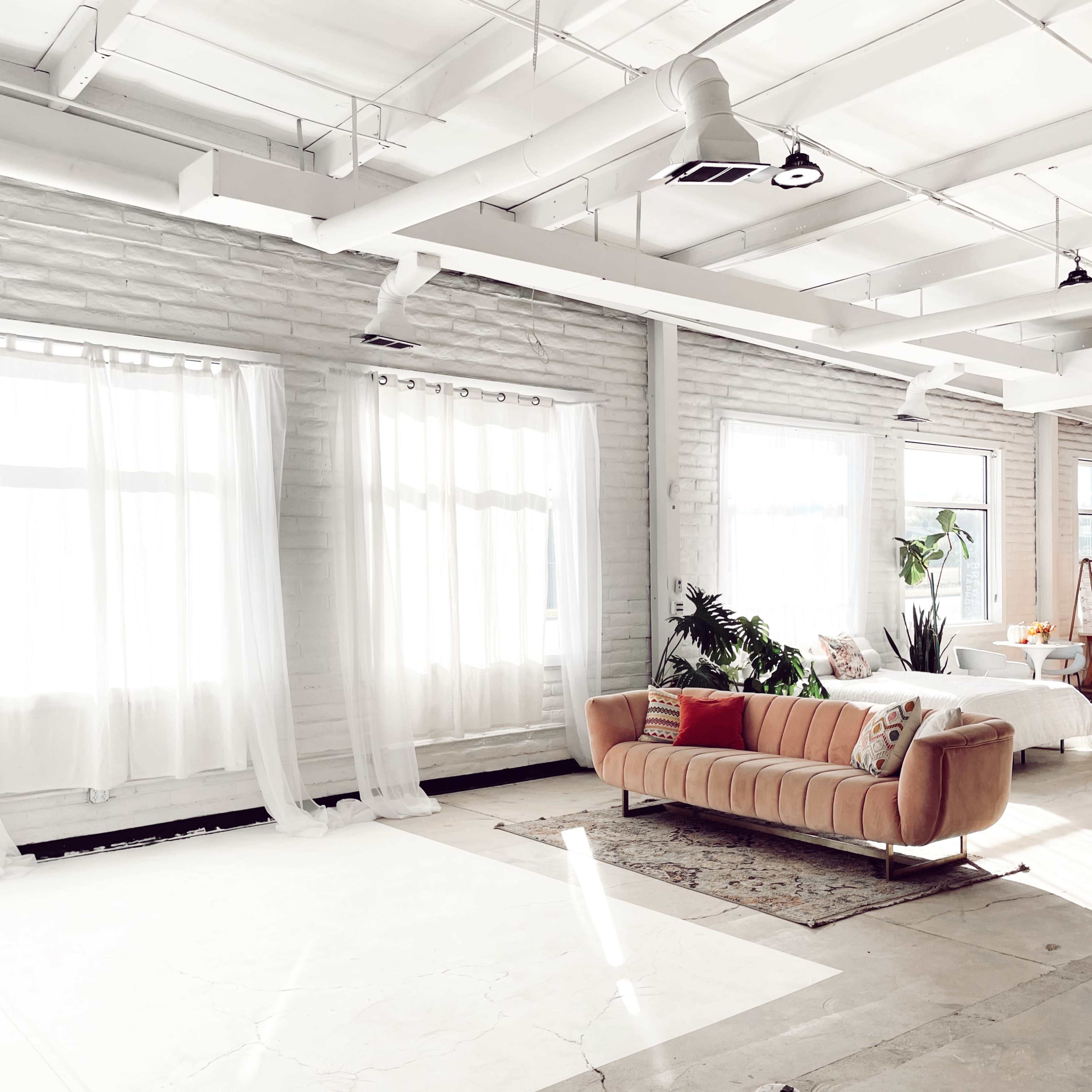 A spacious, well-lit room features a pink couch and large windows with sheer curtains, alongside a bed and plants.