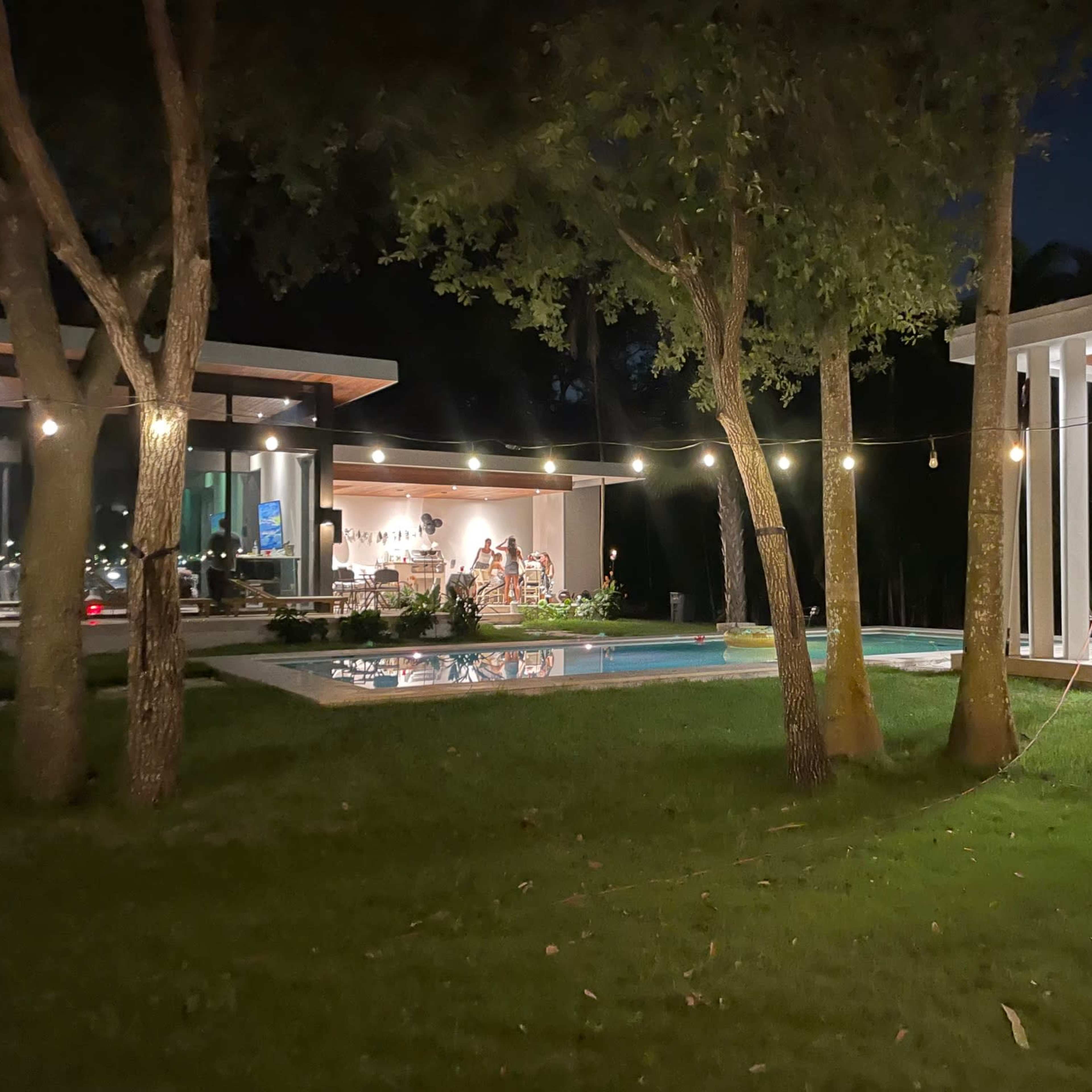 A modern architectural setting at night, featuring a pool surrounded by trees and two distinct structures with warm lights illuminating the area.