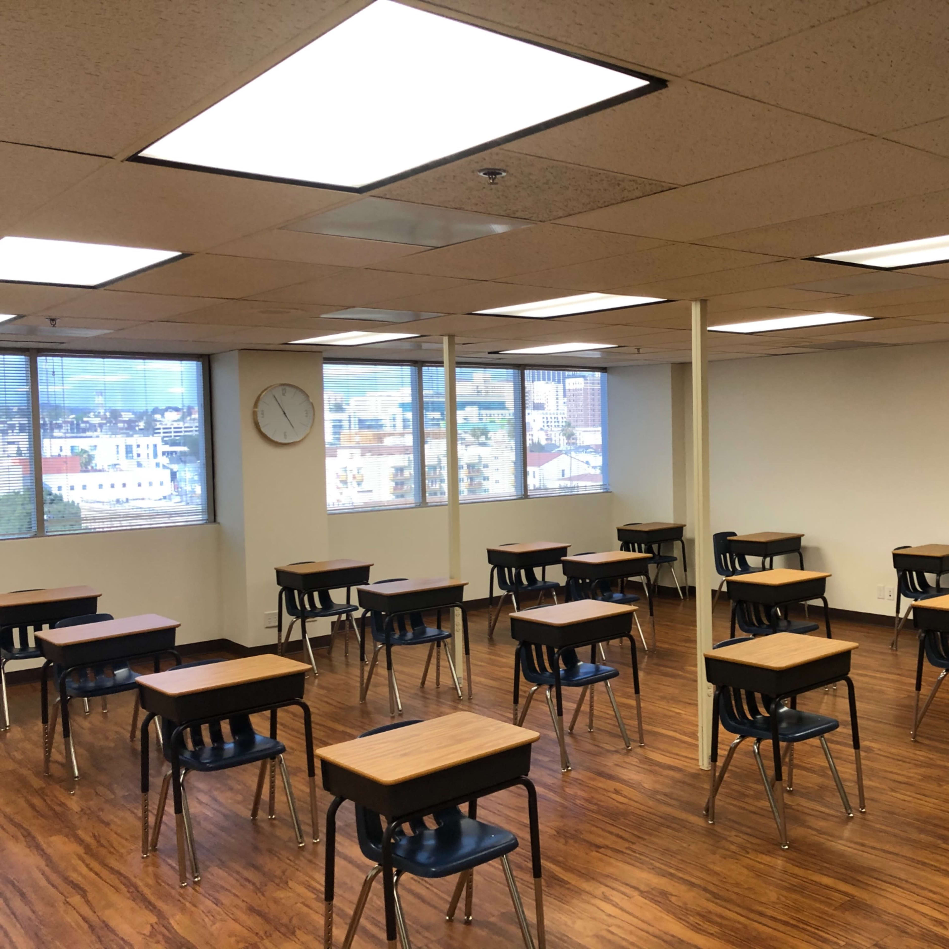 A classroom with wooden desks arranged in rows, large windows providing natural light, and a clock on the wall.
