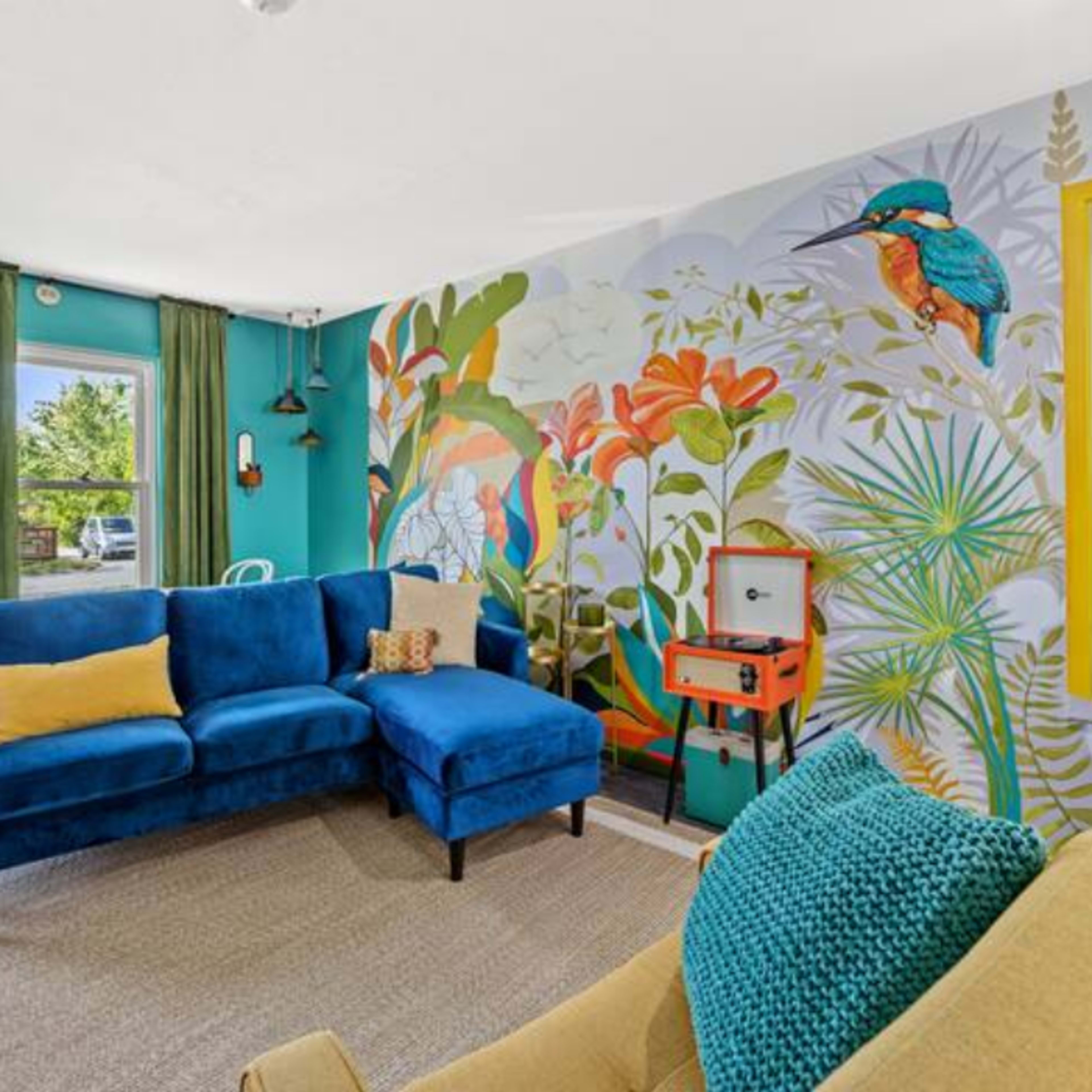 A living room features a blue sectional sofa and a yellow armchair, with a colorful mural of plants and birds on one wall.