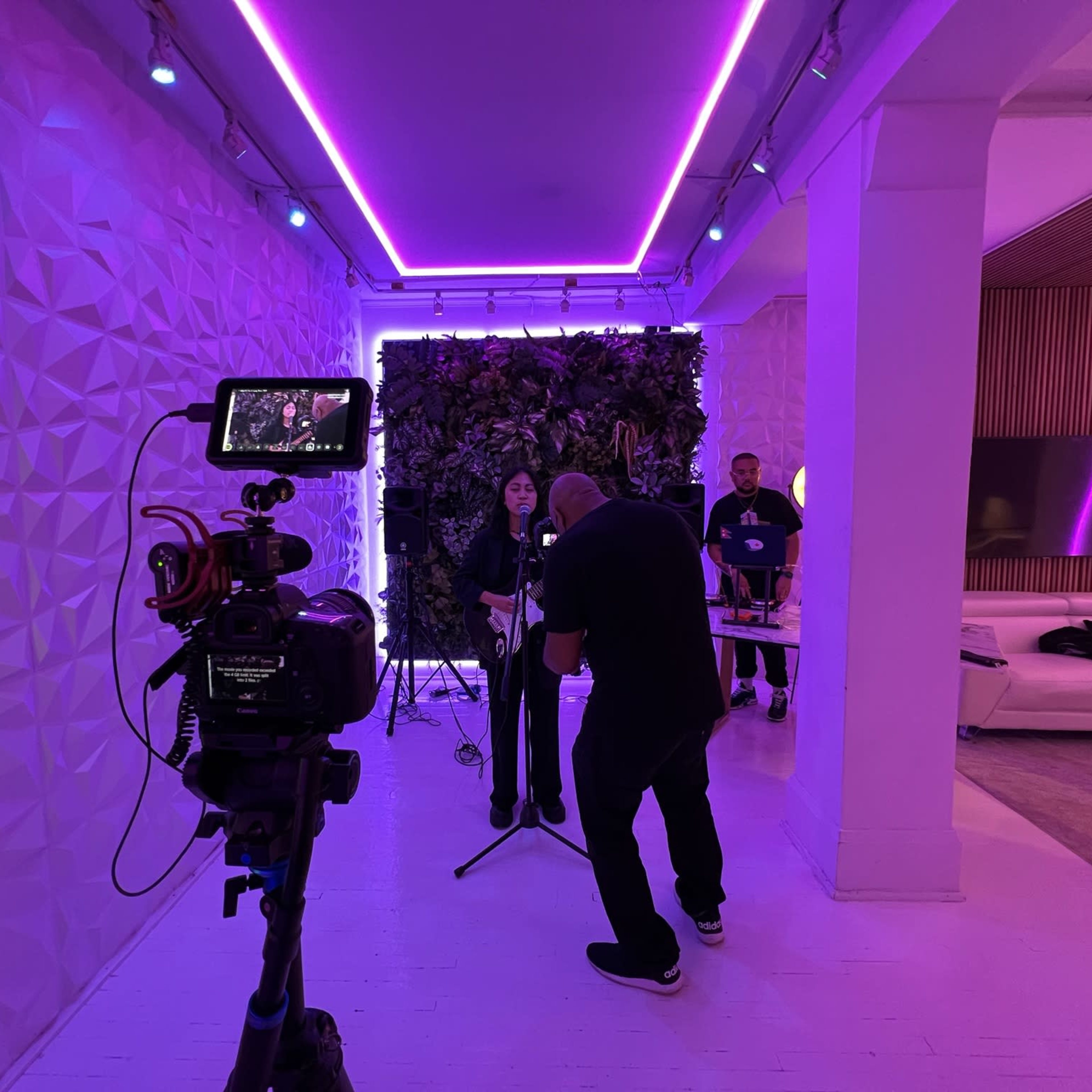 A camera is set up in a brightly lit studio with purple accent lights, capturing a person preparing to perform on a microphone.