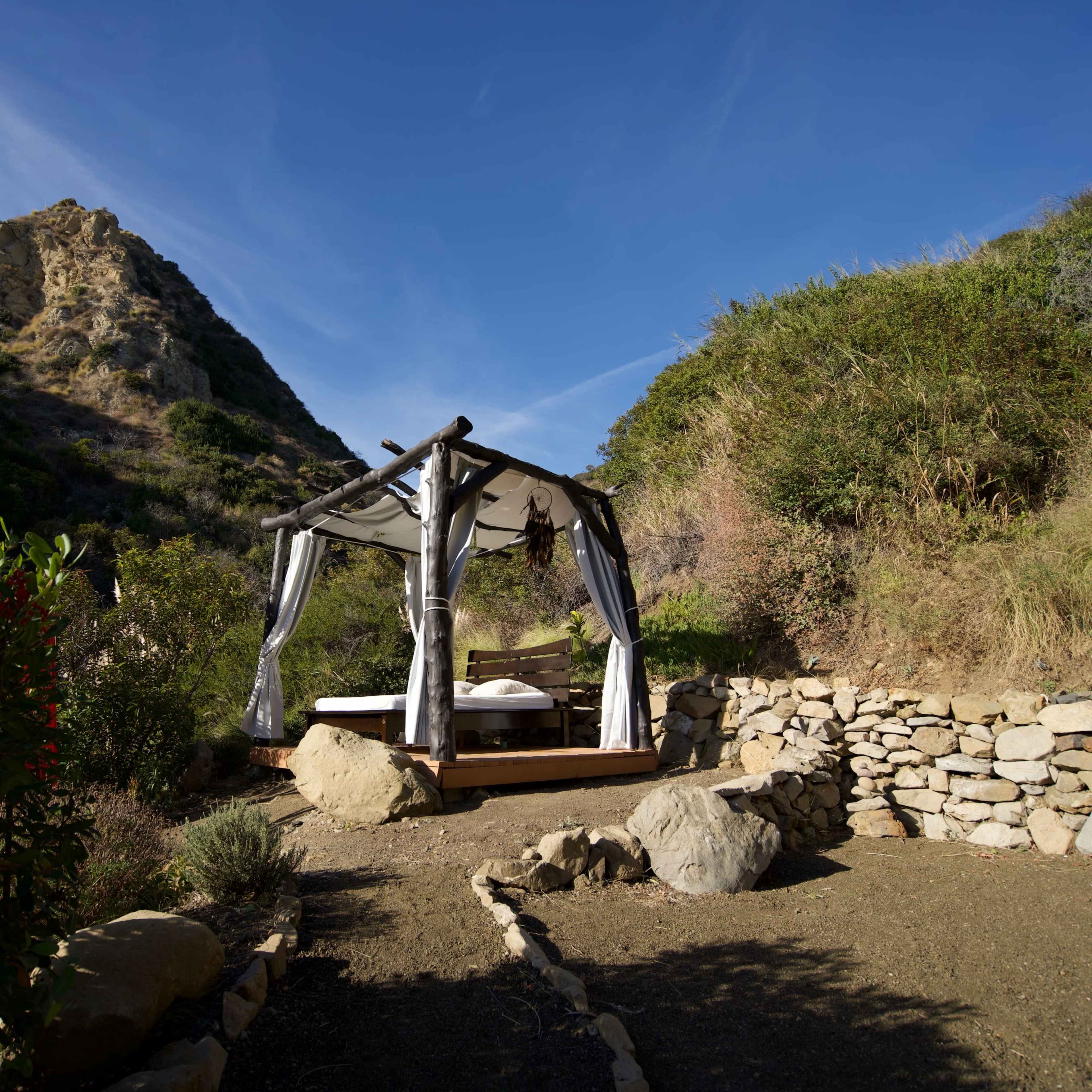 A wooden bed with canopy is situated in a rocky clearing surrounded by bushes, with a mountainous background under a clear blue sky.