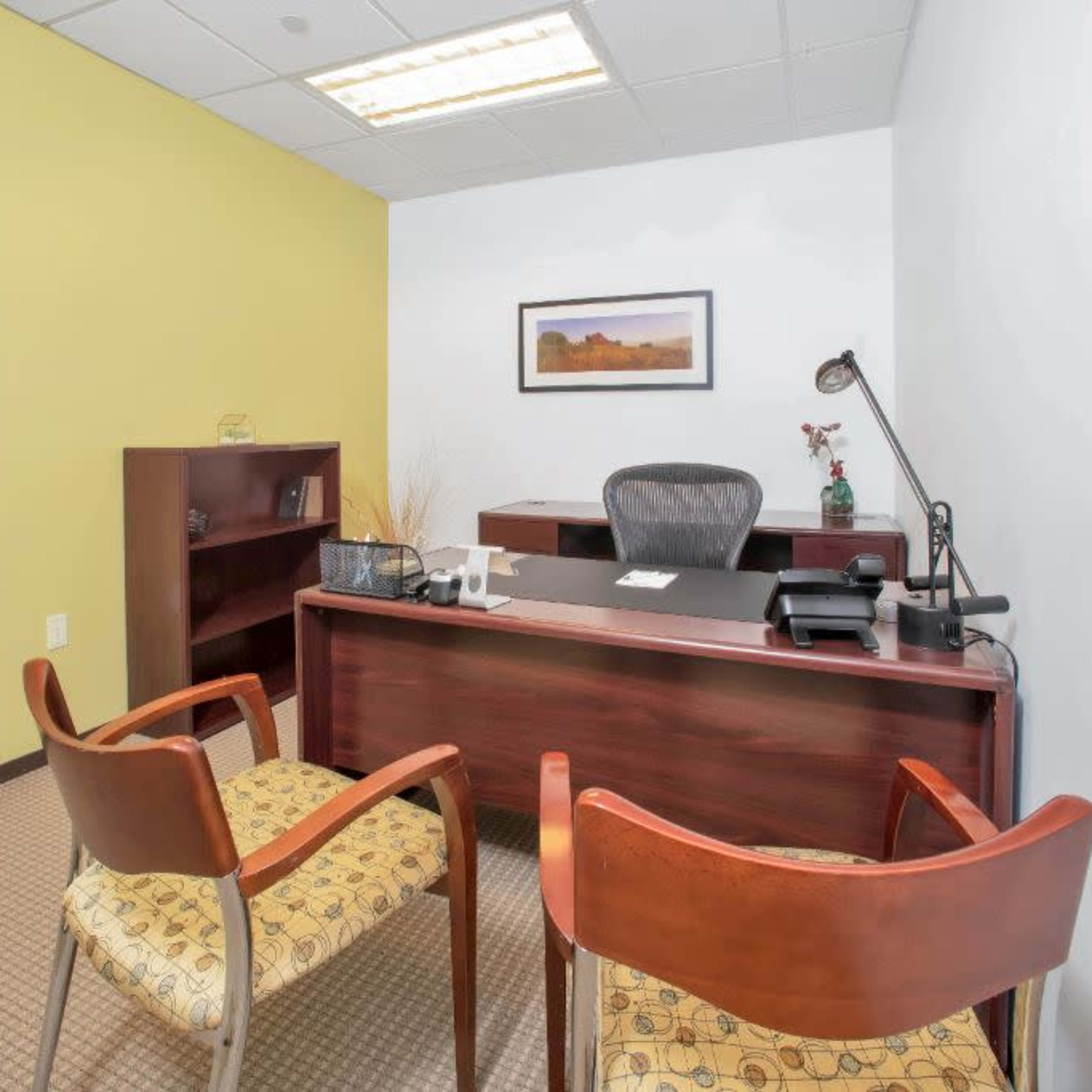 A small office space featuring a desk with a chair, a bookshelf, and two chairs in front of the desk, with a light yellow wall in the background.