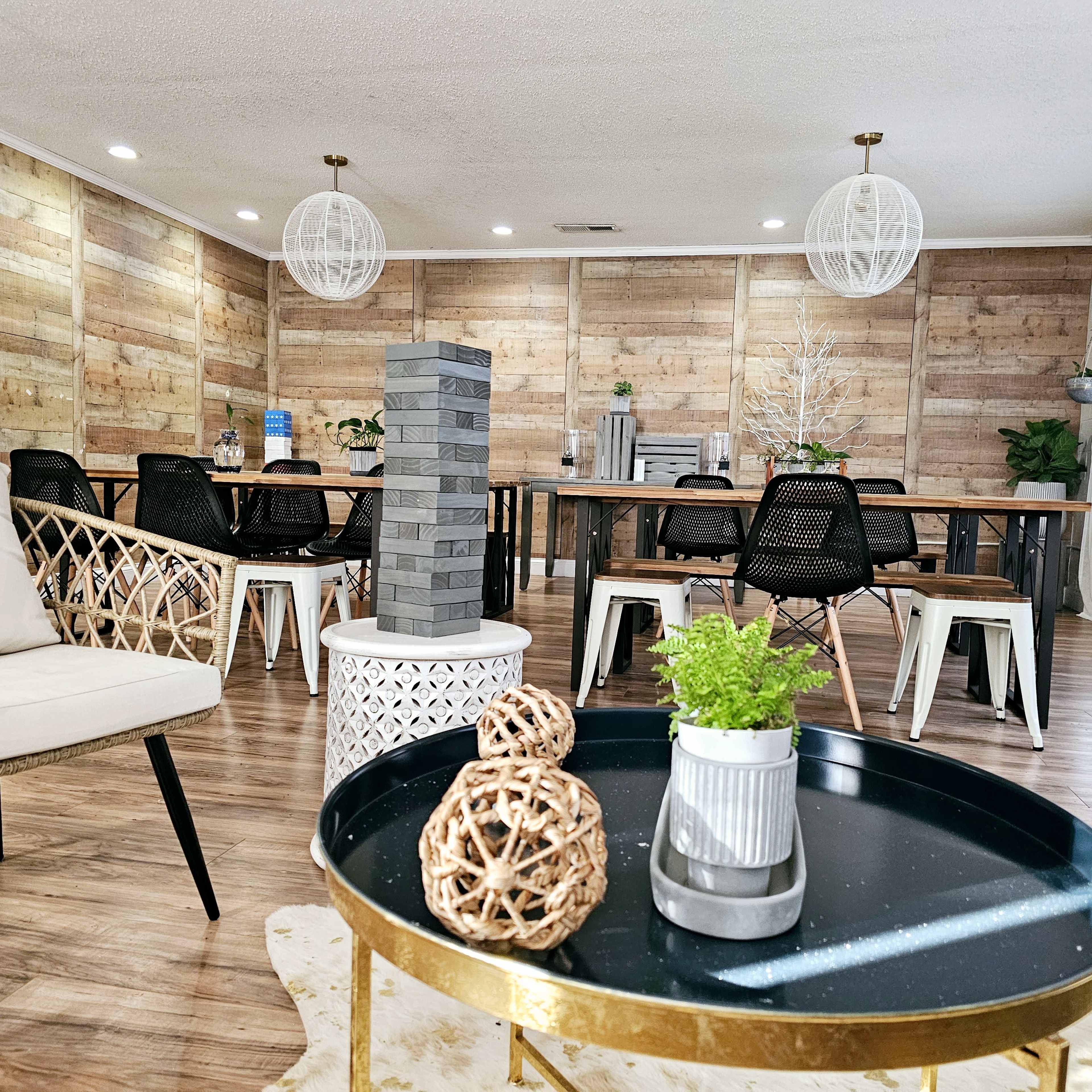 A spacious gathering room with wooden panel walls, a large dining table surrounded by chairs, and decorative elements like plants and a game tower in the foreground.