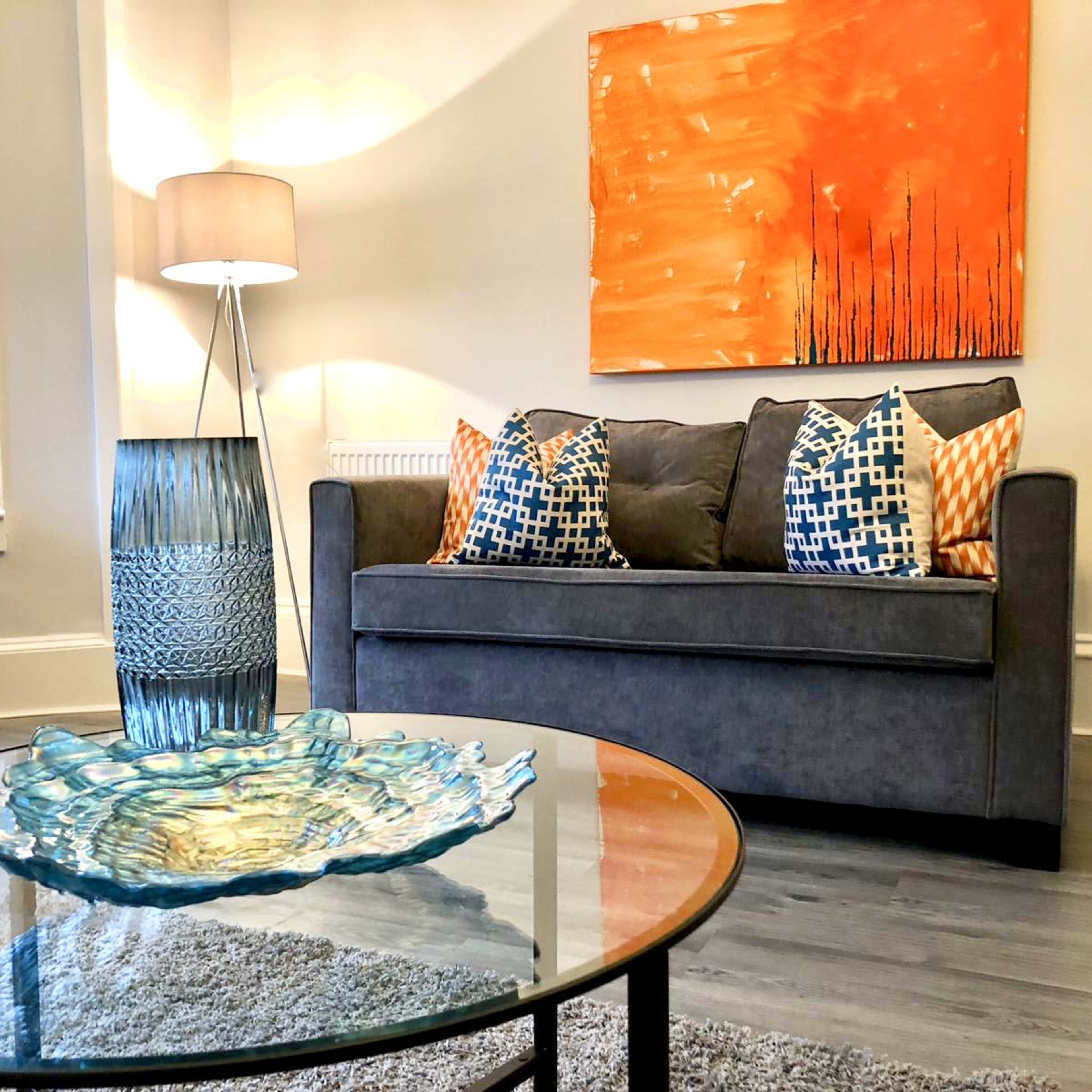 A living room features a gray sofa with decorative pillows, an orange abstract painting on the wall, a glass coffee table, and a blue decorative vase.